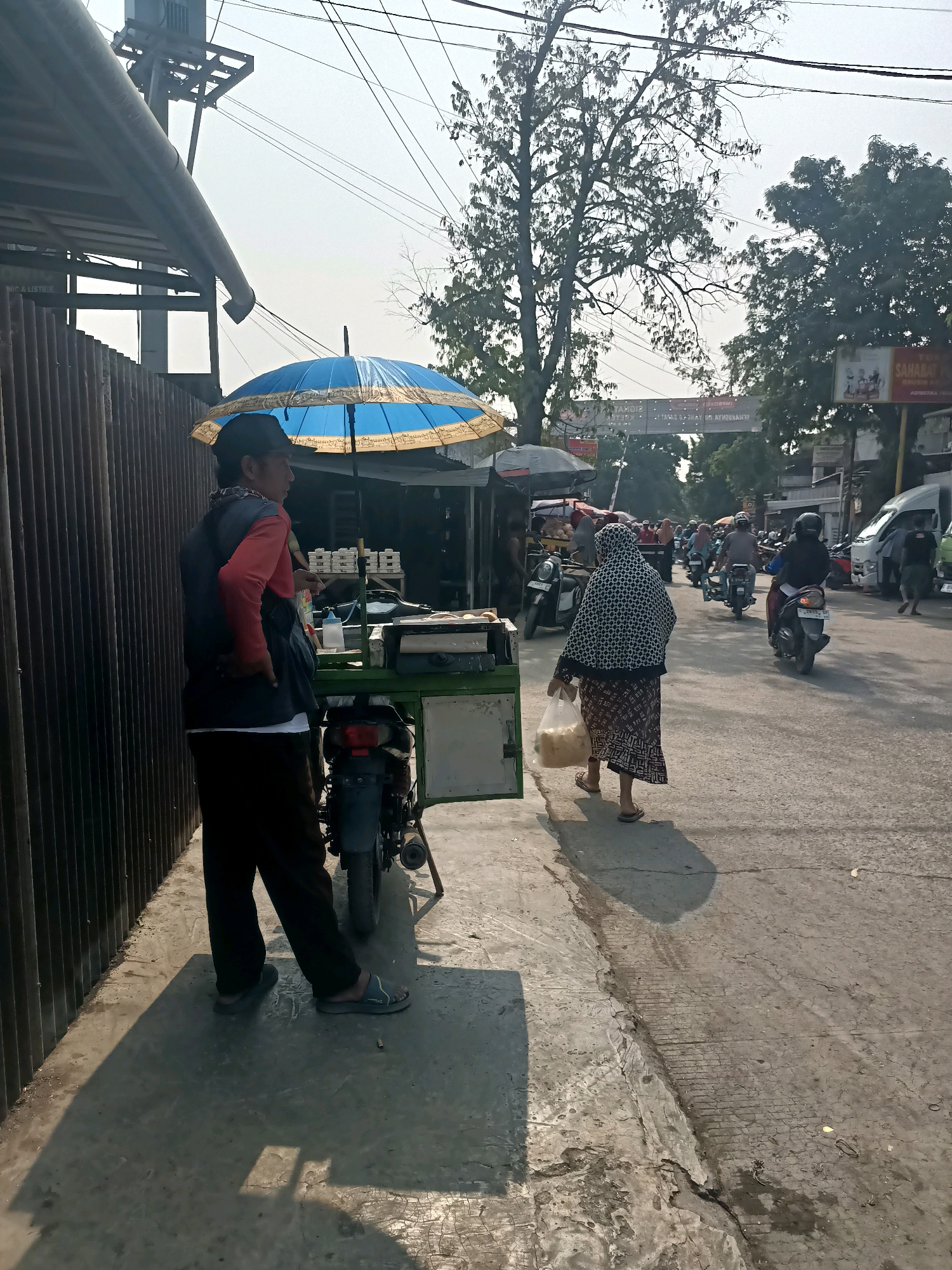 The image depicts a street scene in Ujungrusi, Indonesia, on a sunny day. A man wearing a red long-sleeved shirt, dark vest, dark pants, and a cap stands on the sidewalk, facing right, with his hands on his hips. He is standing next to a food cart covered by a blue and yellow umbrella. The cart has a green frame and holds various items, including what appear to be stacks of food containers. A motorcycle is parked behind the cart.
Further down the street, a woman in a patterned headscarf and dress walks away from the camera, carrying a plastic bag filled with light-colored items. She is wearing sandals. Several motorcycles and a white van are present on the paved road. Trees and utility poles with overhead wires are visible in the background. A sign with text in Indonesian is also visible on the right side of the image. The sidewalk where the man is standing appears to be made of concrete and shows some wear and tear. The overall impression is of a bustling local street with vendors and daily life in progress.