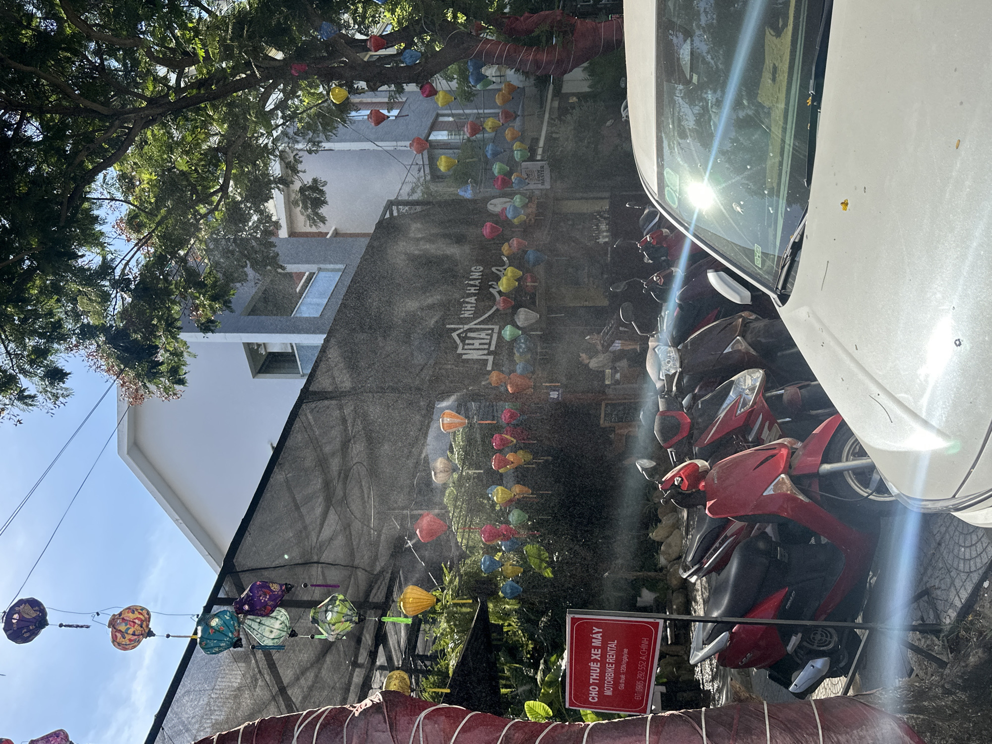 This image, taken on a sunny day in Đà Nẵng, Vietnam, depicts a street scene with a motorbike rental shop. A red sign in the foreground advertises "CHỢ THUÊ XE MÁY MOTORBIKE RENTAL" with pricing information. Behind the sign, several motorbikes are parked, including a prominent red one.
The background features a building with a grey and white facade, covered in part by a dark mesh. Strings of colorful lanterns are suspended above the street, with some hanging from a large tree on the right side of the image. These lanterns are diverse in color and design, including shades of purple, orange, yellow, green, and blue.
A white car is partially visible in the lower right of the frame, its windshield reflecting the bright sun. The street appears to be paved with tiles. The overall impression is a lively and colorful urban environment, typical of many Vietnamese cities.
