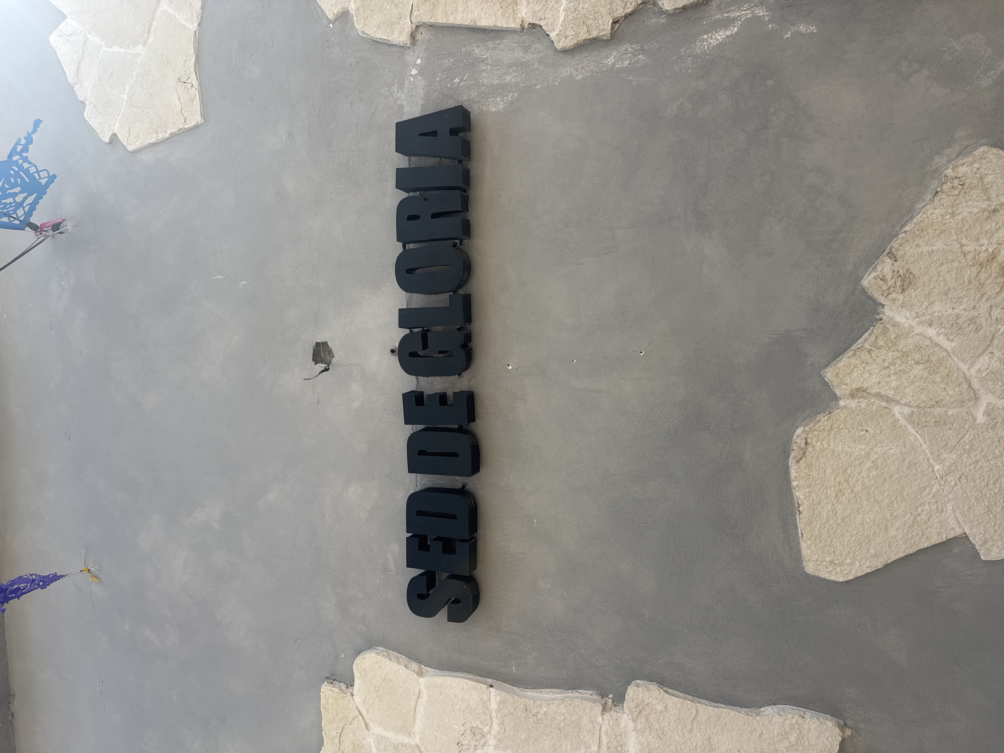 The image is an overhead shot of a gray wall or ceiling with irregular patches of beige stone edging, suggesting a rustic or unfinished aesthetic. The word "SED GLORIA" is vertically spelled out in large, black, block-like letters attached to the gray surface. The location is in Bernal, Mexico. A small piece of blue decorative material is in the upper left corner, and another smaller piece of the same material can be seen in the lower left corner. A small, dark hole with protruding wires is visible to the left of the word.