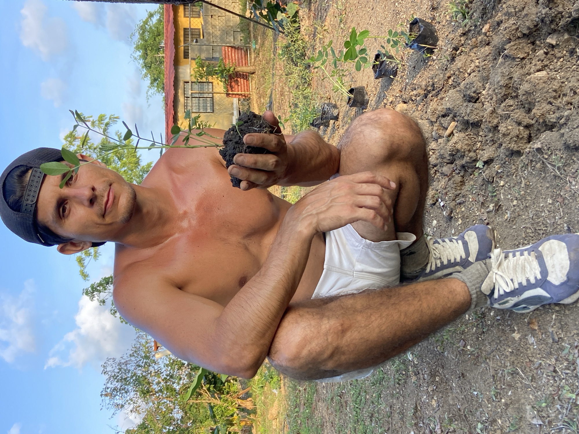 A man with dark hair and a black baseball cap is crouched in a dirt area, holding a seedling with roots in his right hand. He is shirtless, wearing white shorts and athletic shoes. The background shows a yellow building with windows, a chain-link fence, and trees under a partly cloudy sky. Several other seedlings in small black pots are visible in the foreground and mid-ground. The scene appears to be outdoors, possibly a garden or yard, in the city of Los Velásquez, Venezuela.