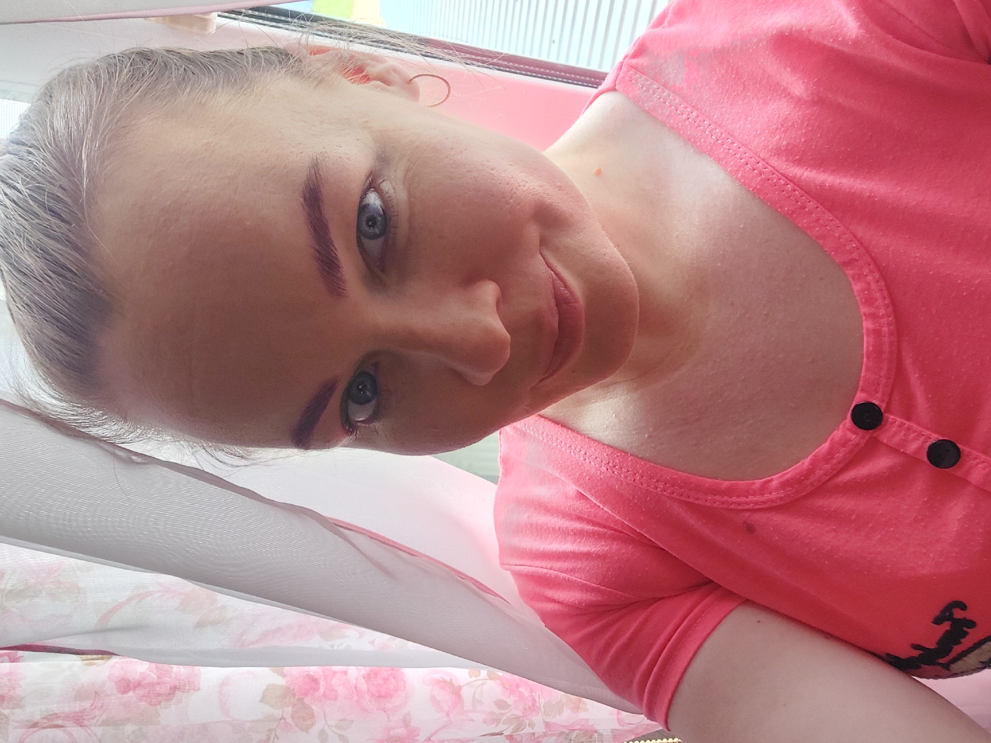 The image is a close-up, slightly angled selfie of a woman's face and upper torso, taken indoors. She has fair skin with visible pores and some redness. Her hair is pulled back into a bun, and she has light-colored eyebrows. She is wearing a bright pink t-shirt with two black buttons at the neckline. She has a small hoop earring in her visible right ear and a small, light-colored mole on her neck. The background shows pink and white floral curtains and a window with horizontal blinds. The overall lighting is bright, suggesting it is daytime. The image does not contain any discernible information to place it in the city of Большой Букор, Russia.