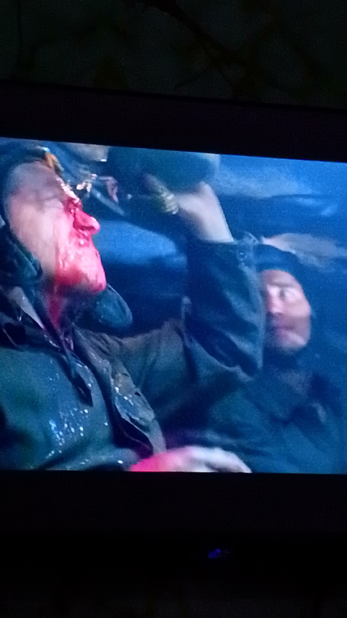 The image shows two men in what appears to be a dark, confined space. One man is heavily bloodied around the face, particularly the nose. Both men are wearing dark-colored clothing with visible textured material. The environment is dimly lit and has a bluish hue. The location is not discernible from the image. The action appears to be related to conflict or injury.