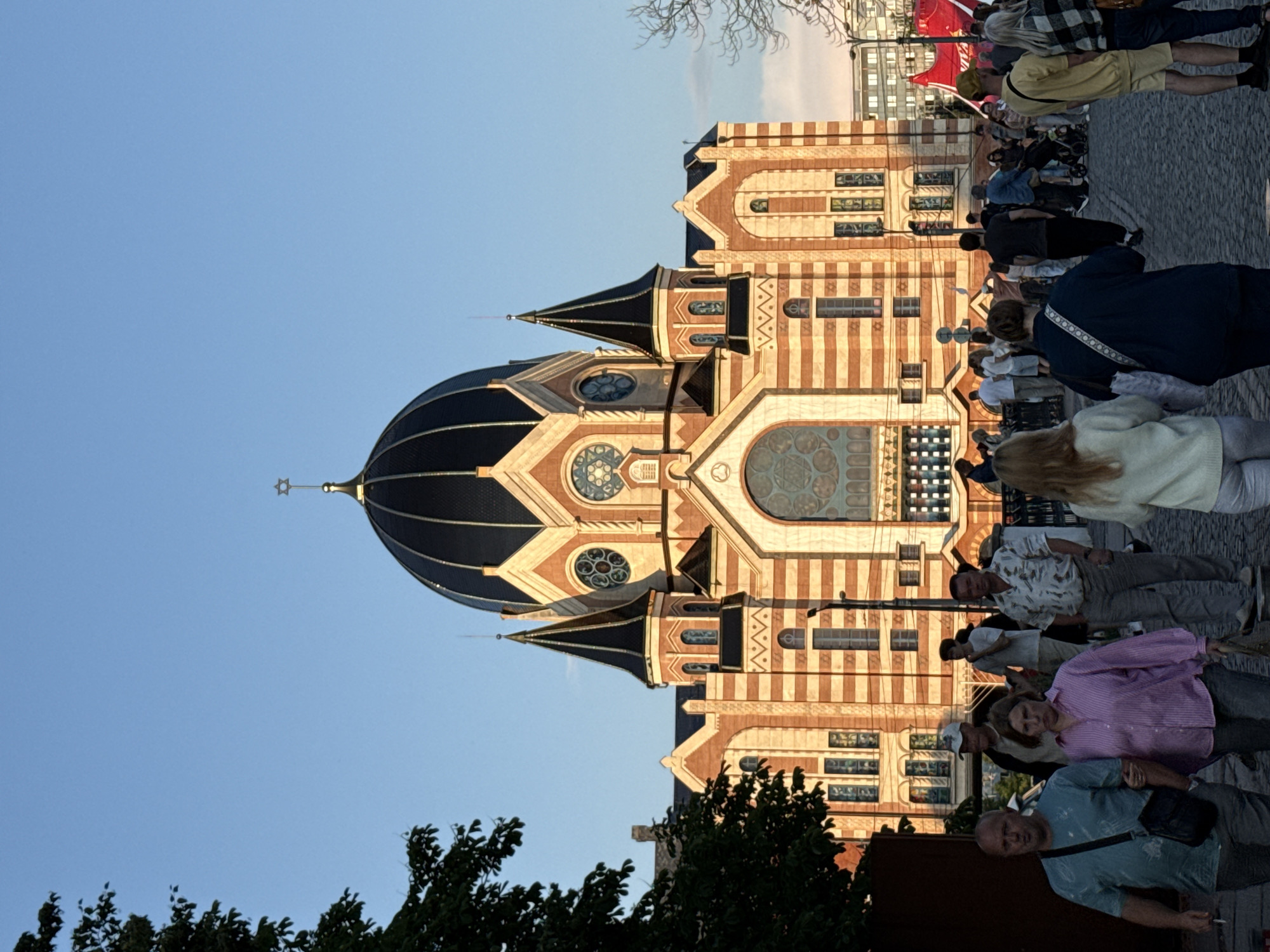 The image shows the New Synagogue in Kaliningrad, Russia. The building features a large, dark dome topped with a Star of David, flanked by smaller spires. The facade is constructed of alternating bands of red brick and lighter stone. A crowd of people are walking on a cobblestone street in front of the synagogue, some arm in arm. The sky is a clear blue, indicating daytime. The sun appears to be setting or rising, casting a warm light on the building.