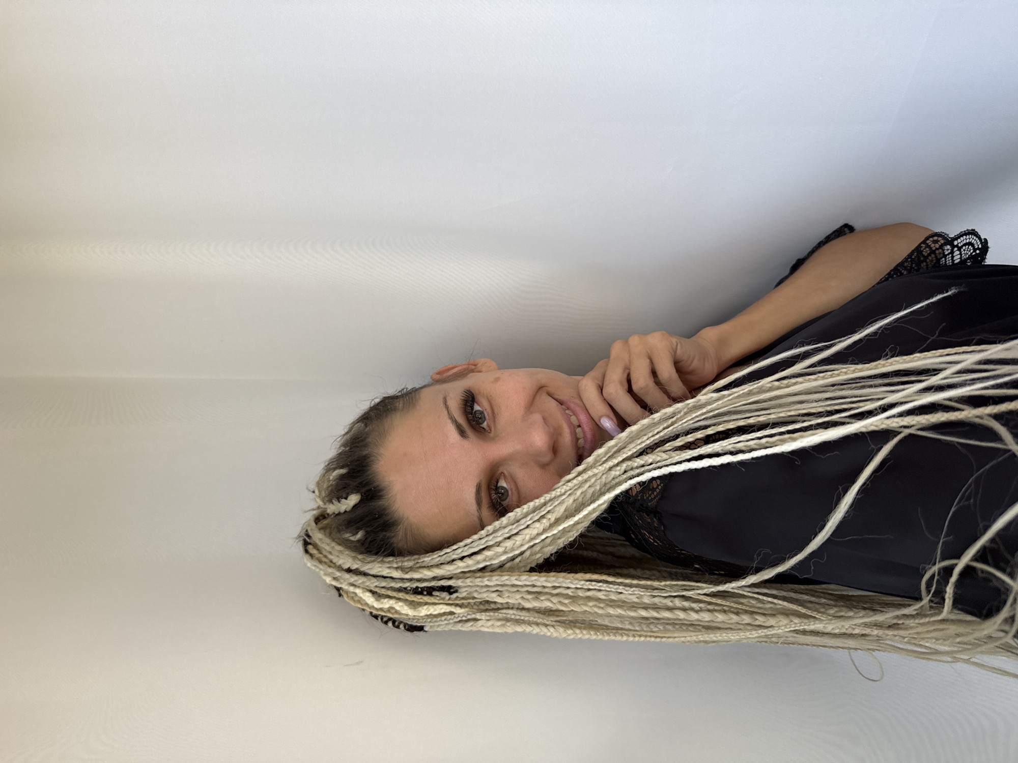 A woman with blonde braided hair stands in a corner formed by two white walls. She is wearing a black top with lace trim. Her right hand is raised to her chin, and she is looking at the camera with a slight smile. The lighting suggests an indoor environment, likely a home or studio, with soft natural light coming from an unseen source to her left, casting subtle shadows on the wall. The image is a portrait, focusing on the woman's face and upper body, with the background providing a neutral setting. The specific location of Rusanovo, Russia, is not determinable from the visual information present in the image.