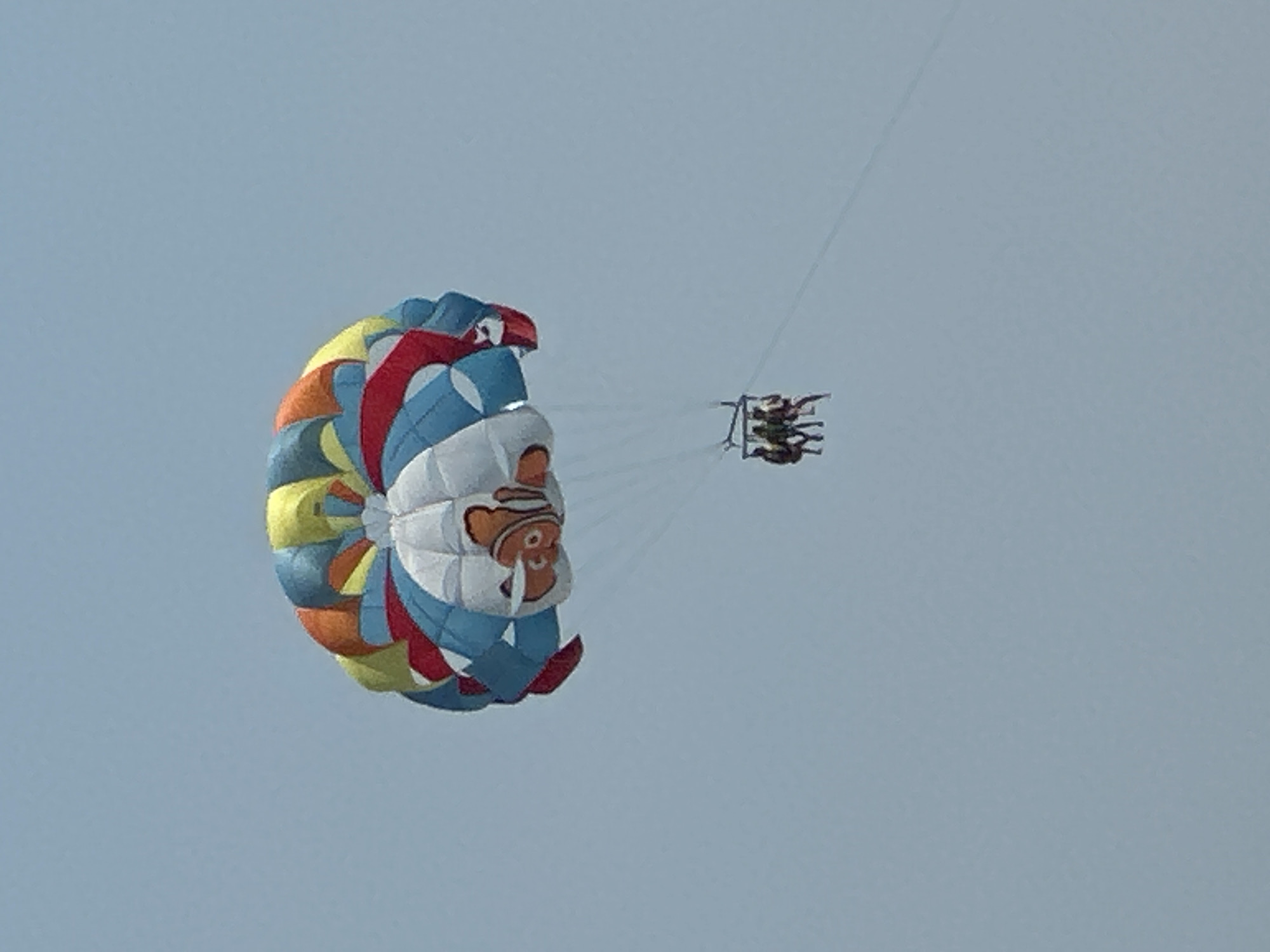 In a bright, clear sky, a parasail adorned with a cartoon clown face is prominently featured. The parasail, a vibrant mix of red, blue, yellow, and orange sections, displays the smiling face of a clown, complete with rosy cheeks and a striped hat. Below the parasail, a small raft carrying at least three people is visible, suspended by thin lines. The raft itself is dark and appears to be a recreational vessel. The overall scene suggests an outdoor recreational activity, likely taking place on a sunny day with good visibility. The vast expanse of the pale blue sky serves as the backdrop, with no other discernible landscape features. The image captures a moment of leisure and adventure.