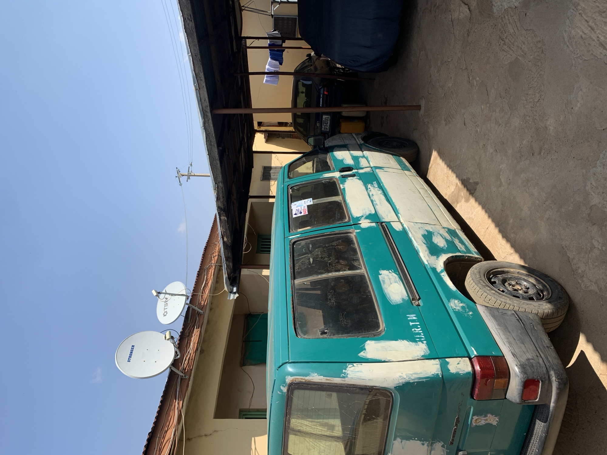 The image shows a teal van parked in a lot in Rafin Par, Nigeria. The van has white putty covering areas of the body, indicating it is undergoing repairs. Next to the van, partially obscured by a dark blue cover, is a dark-colored car. Clothes are hanging from a line under the overhang of a roof. On the wall of a nearby building are two satellite dishes. The sky is clear and blue.