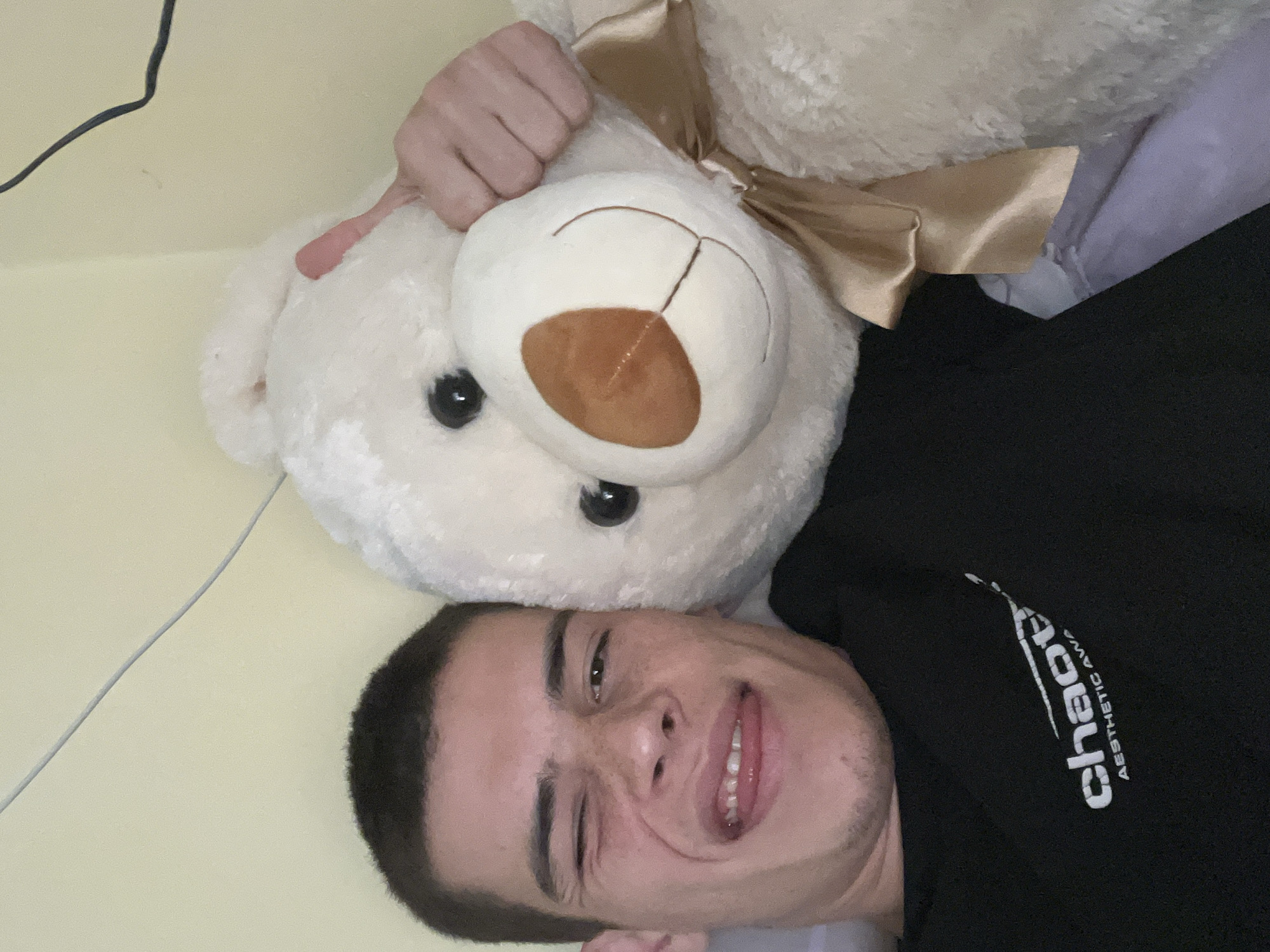 A selfie shows a person with short dark hair and light skin, wearing a black shirt with the word "Chaos" printed in white, lying on their back with a white teddy bear positioned above their head. The teddy bear has a brown nose and a gold ribbon tied around its neck. A black cable is visible in the background. The location cannot be determined from the image.