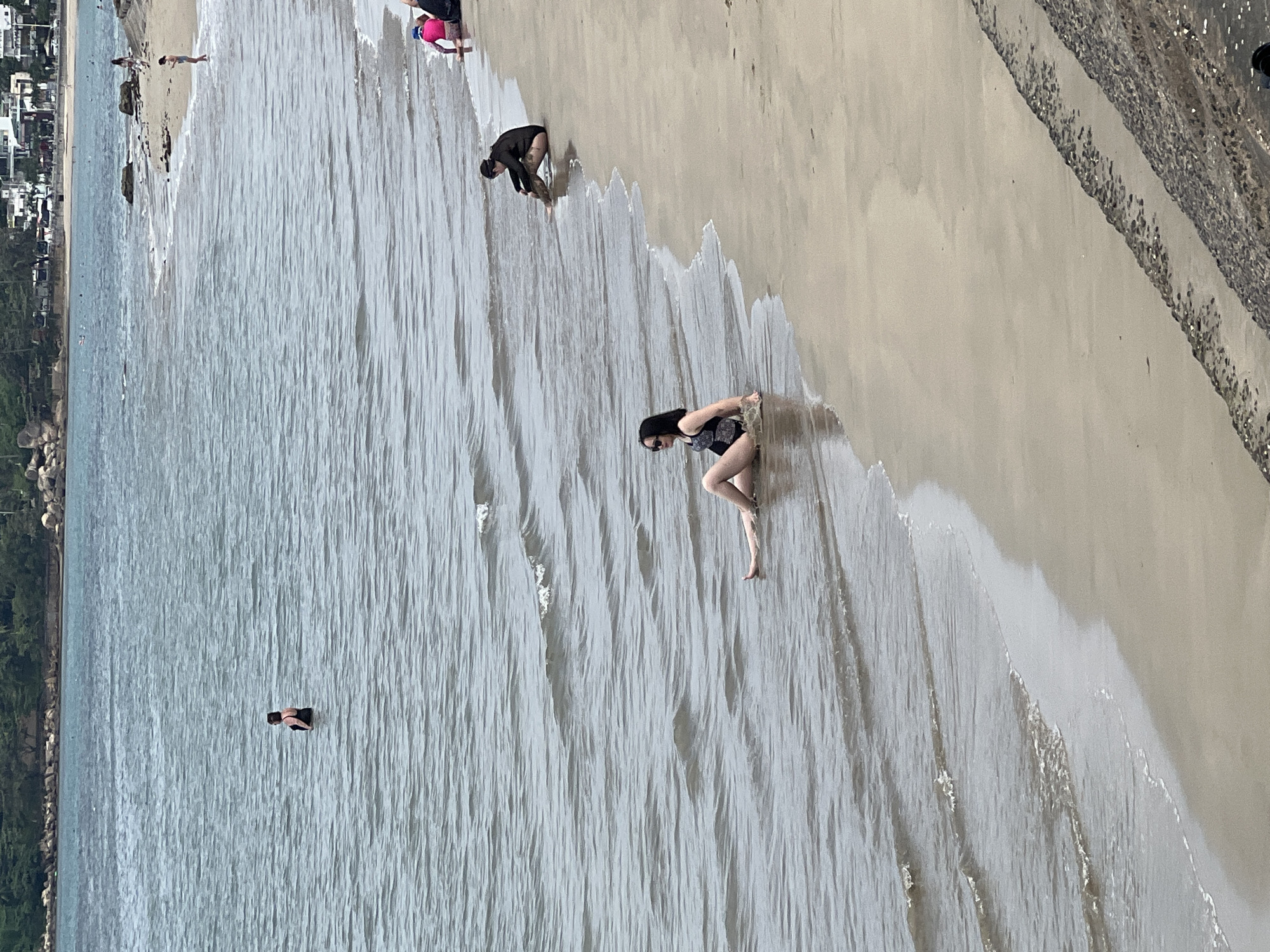 The image captures a vertically oriented view of a bustling beach scene, likely from an elevated perspective. The primary focus is on the interface between the light beige sand and the calm, blue-grey ocean water, which shows gentle, receding waves.
In the mid-ground, a woman wearing a dark, patterned swimsuit is kneeling at the water's edge on the sand, with her hands in the shallow water. Further to the left and slightly behind her, another person, dressed in a dark long-sleeved top and dark bottoms, is also bent over near the waterline, appearing to interact with the sand or water. Closer to the background, on the sandy beach, a person is partially visible next to a bright pink object, possibly a bag or piece of clothing. To the far left, a person in dark swimwear is standing chest-deep in the ocean, facing away from the viewer. Several other indistinct figures can be seen further down the beach and in the distance.
The background reveals a distant coastline featuring a mix of buildings and lush green vegetation, suggesting an urban or semi-urban setting. A textured, dark wall or barrier runs along the far right edge of the beach. The scene takes place during bright daylight, with no strong shadows, indicating sunny weather. The overall impression is one of a leisure activity at a coastal location, consistent with Bac Nha Trang Ward, Vietnam. No visible text is present in the image.