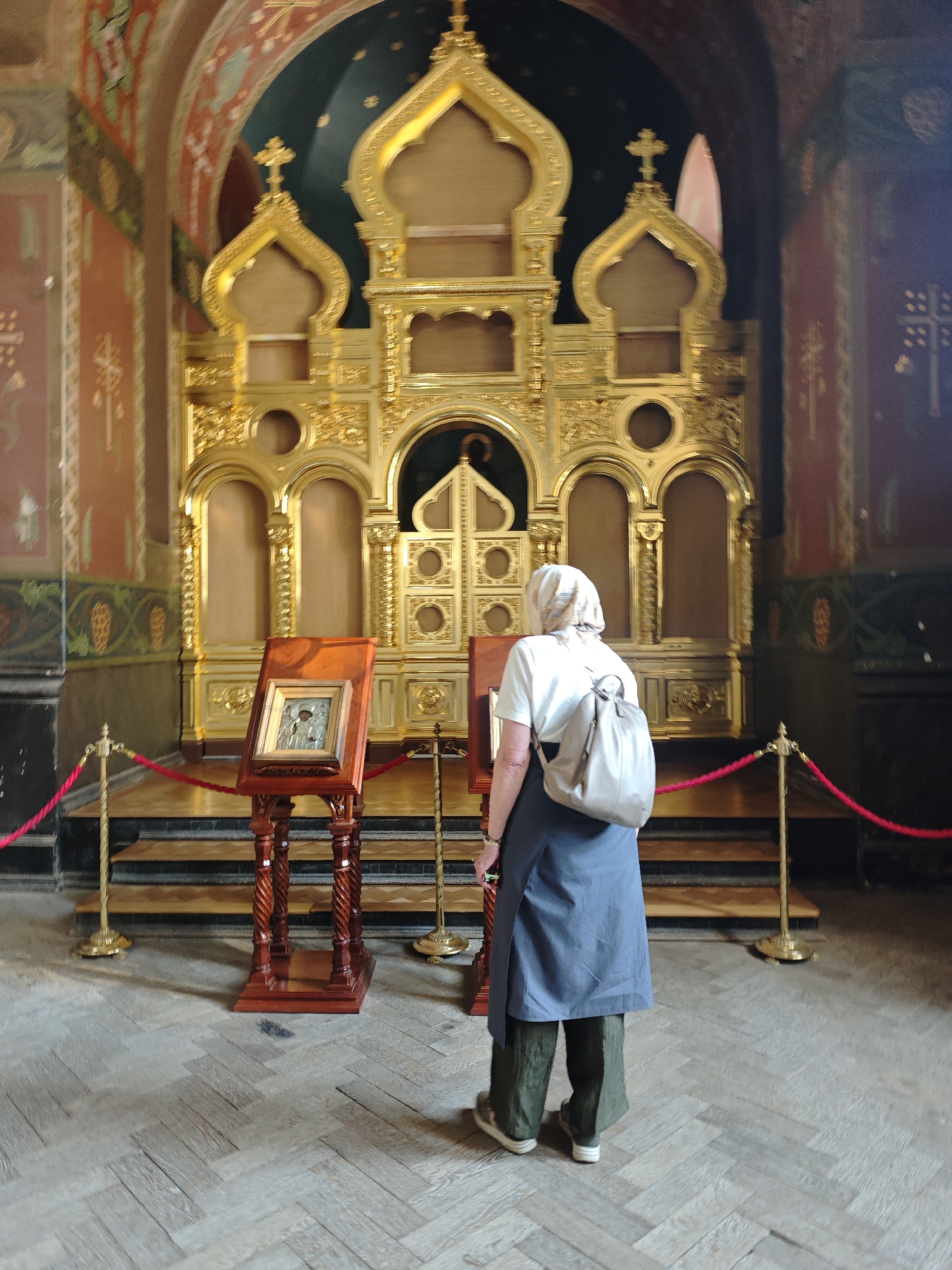 An eye-level indoor shot displays the interior of an ornate Eastern Orthodox religious building in Unknown, Unknown. A prominent, multi-tiered golden iconostasis with intricate carvings, arched sections, and several onion dome-shaped elements topped with Orthodox crosses dominates the background. The central Royal Doors are visible, slightly ajar. In the mid-ground, a dark wooden analogion (lectern) with carved legs holds a framed icon depicting a religious scene or figure in metallic relief. A red velvet rope, suspended between two golden stanchions, acts as a barrier in front of the iconostasis. An adult female, viewed from behind, stands to the left of the frame, facing the iconostasis and the displayed icon. She is wearing a light-colored headscarf, a white short-sleeved top, a long dark blue/grey tunic over dark olive-green wide-leg trousers, and light-colored flat shoes, carrying a beige backpack. The floor is light-colored wood laid in a herringbone pattern. The walls feature painted murals with religious patterns in red, green, and gold tones. Dark wooden steps lead up to the area directly in front of the iconostasis.