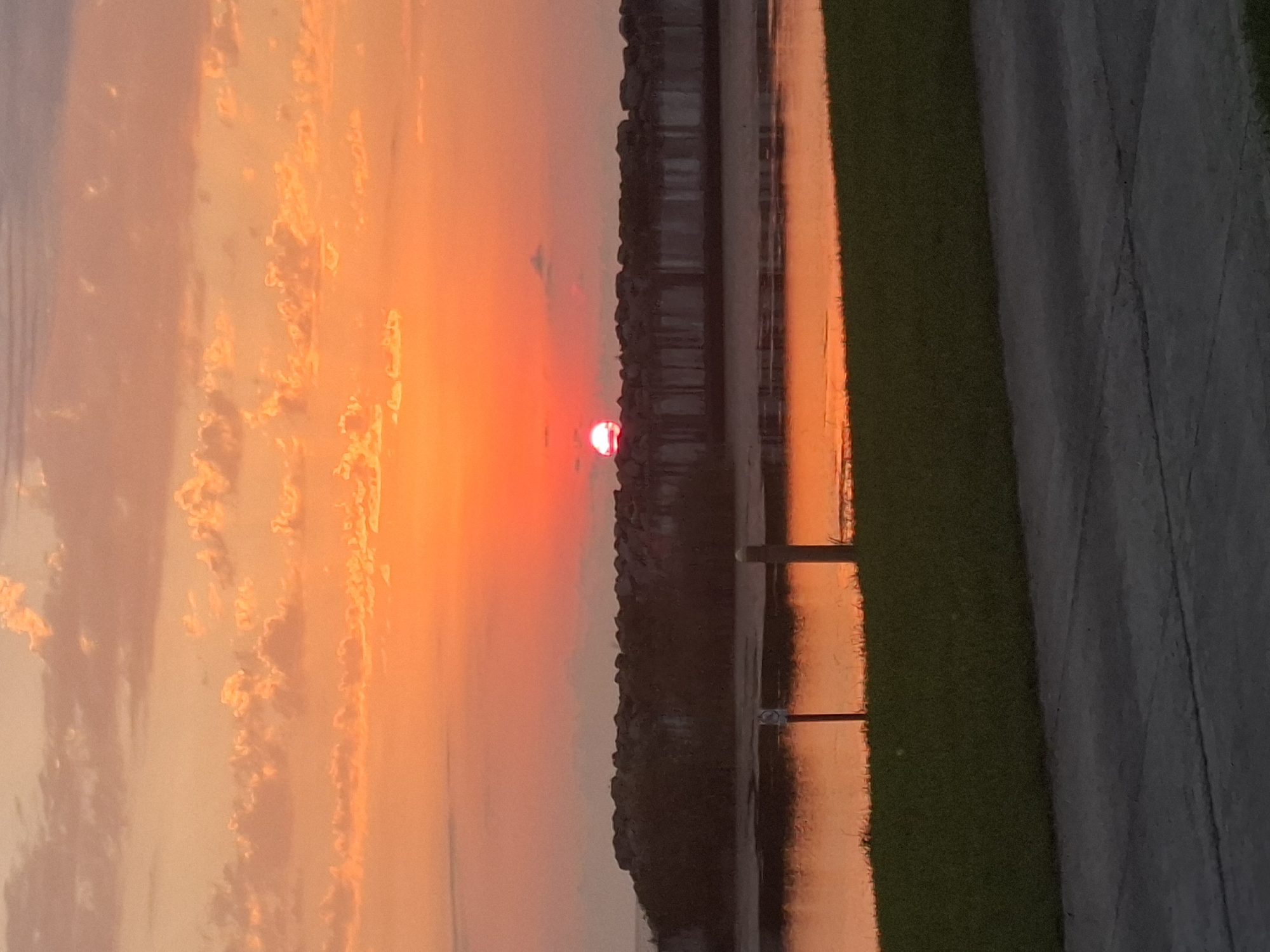 The image captures a sunset over the Black Sea in Anaklia, Georgia. The sky is ablaze with vibrant orange and pink hues, with scattered clouds tinged with the same warm colors. The sun, a bright red orb, is partially obscured by a line of dark, rocky structures that appear to be a breakwater or pier. Below the structures, a calm body of water reflects the colors of the sky. In the foreground, a strip of green grass meets a gray concrete pathway. There are no people or discernible animals in the image.