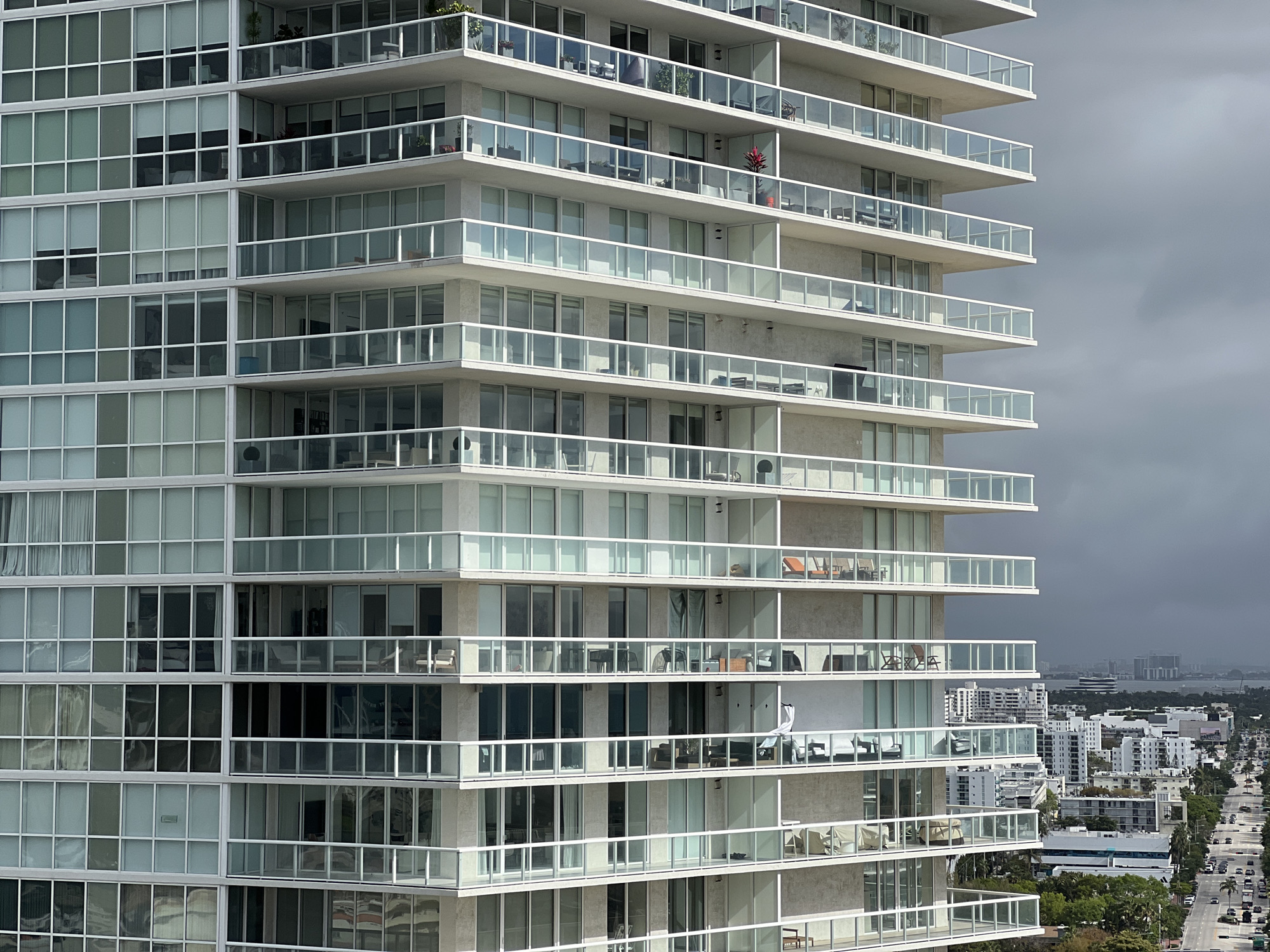 The image shows a modern, high-rise condominium building in Miami Beach, Florida, USA. The building's architecture features multiple floors with projecting balconies enclosed by glass railings. Each floor has numerous windows, reflecting the overcast sky. The building is made of white concrete. In the background, a cityscape is visible with other high-rise buildings and a street lined with cars and trees. The sky is overcast.