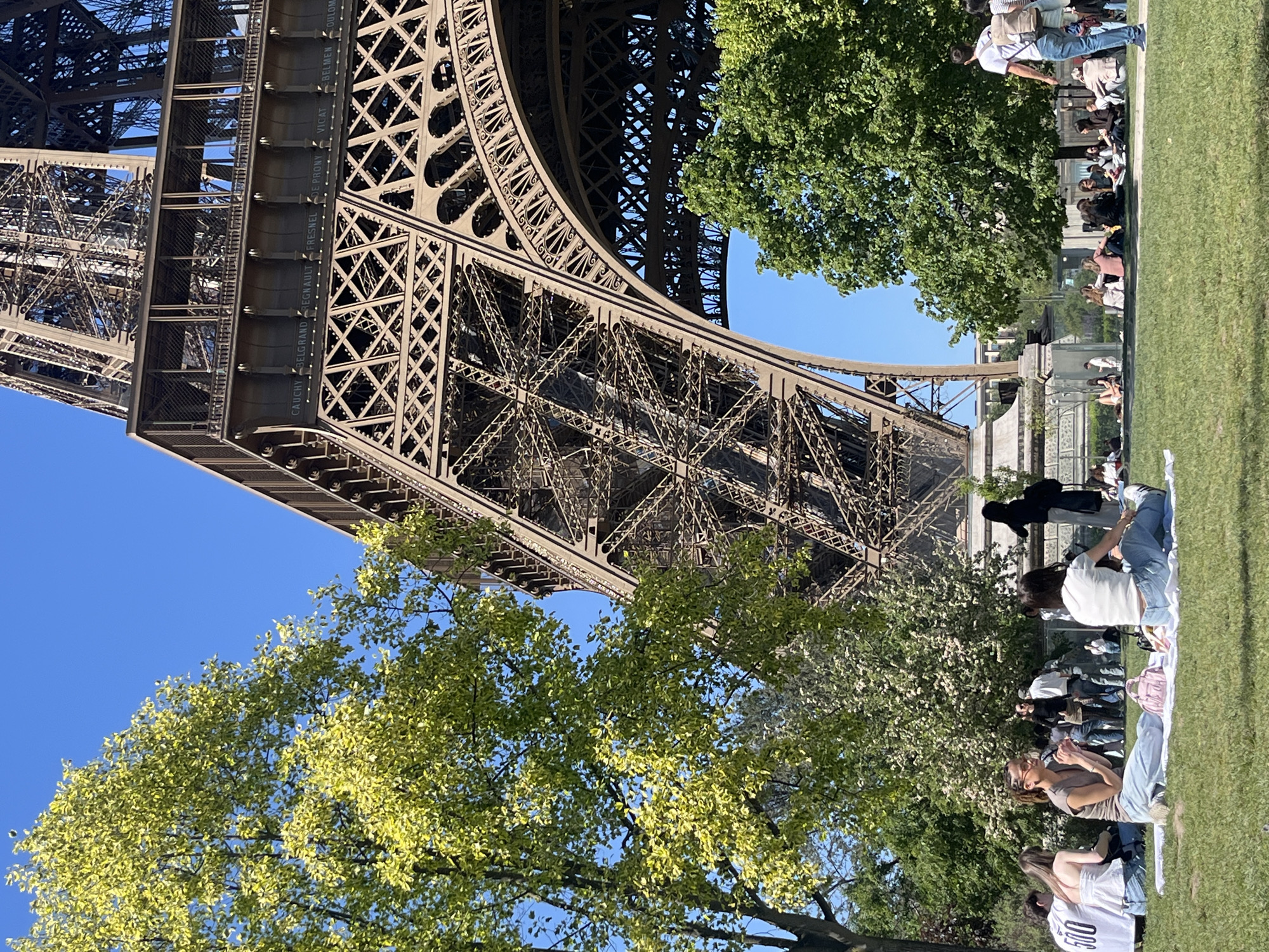The image shows the Eiffel Tower in Paris, France, from a low angle. The lower portion of the tower is visible, showcasing its intricate metal lattice structure. The sky is clear blue. Some green trees partially obscure the base of the tower. On the right side of the image, a group of people is sitting on the grass. Some individuals are wearing casual clothing, and some are looking at the tower. The ground appears to be a grassy area.