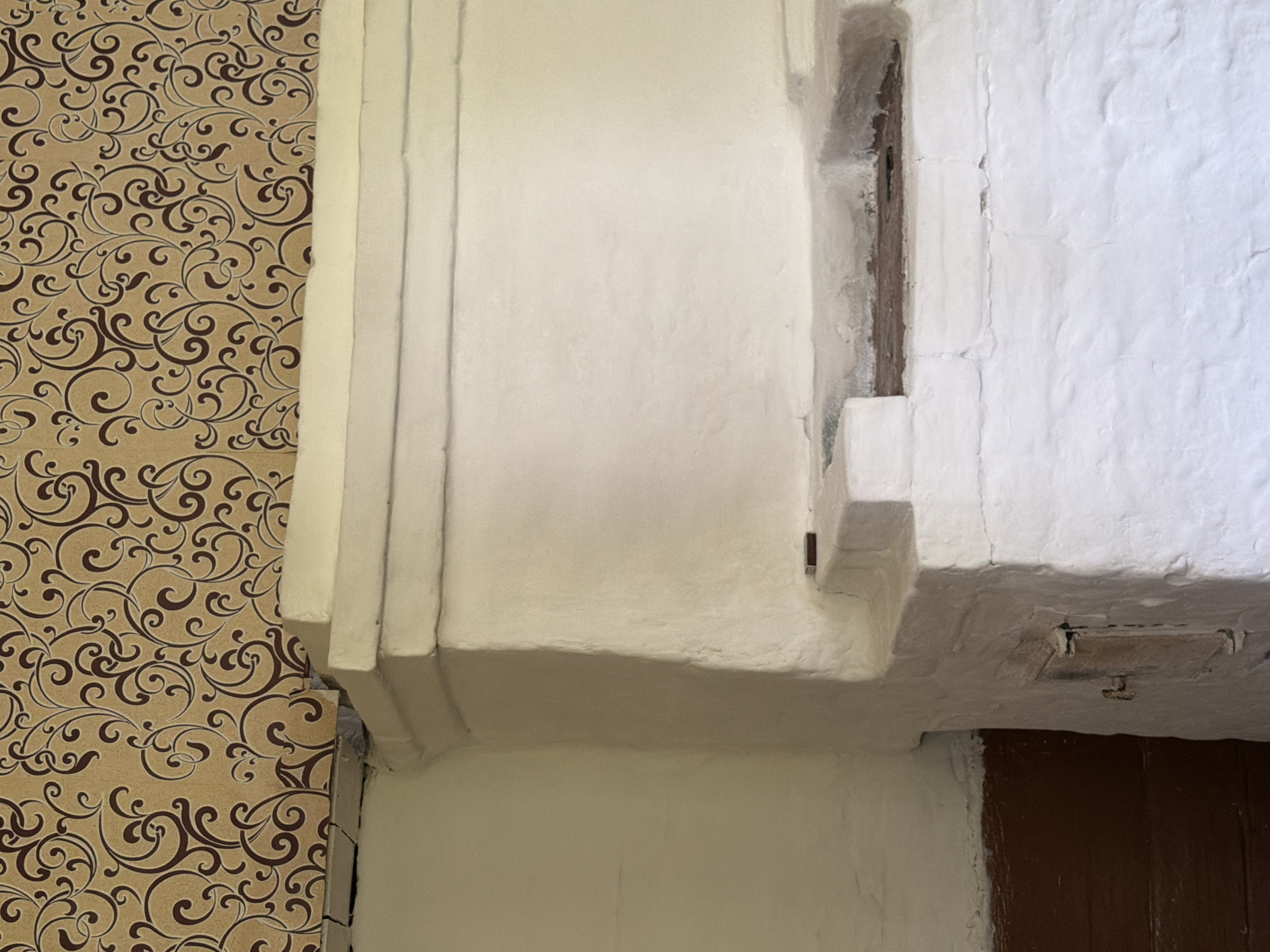 The image shows the interior of a rustic room. The dominant feature is a large, white, plastered stove, likely a traditional Russian stove (pech). The stove has a rough texture, suggesting it is made of brick or clay and has been whitewashed. There are visible cracks and imperfections in the plaster. The stove has a rectangular opening near the bottom, likely a firebox with a metal door, and a horizontal shelf-like structure above it, possibly for heating or cooking.
The wall above the stove is covered with wallpaper featuring an intricate, swirling pattern in brown and beige. The wallpaper appears to be older, with some slight damage visible near the corner where it meets the stove.
To the left of the stove, a portion of the wall is visible, also plastered and painted white, with a darker wooden paneling at the bottom, possibly wainscoting.
The overall impression is one of a simple, functional living space, likely in a rural or older dwelling. The scene is static and does not contain any people or immediate action. The lighting suggests an indoor environment, possibly with natural light coming from an unseen source. The specific location in Kameshki, Russia, implies a context of traditional Russian architecture and domestic life.