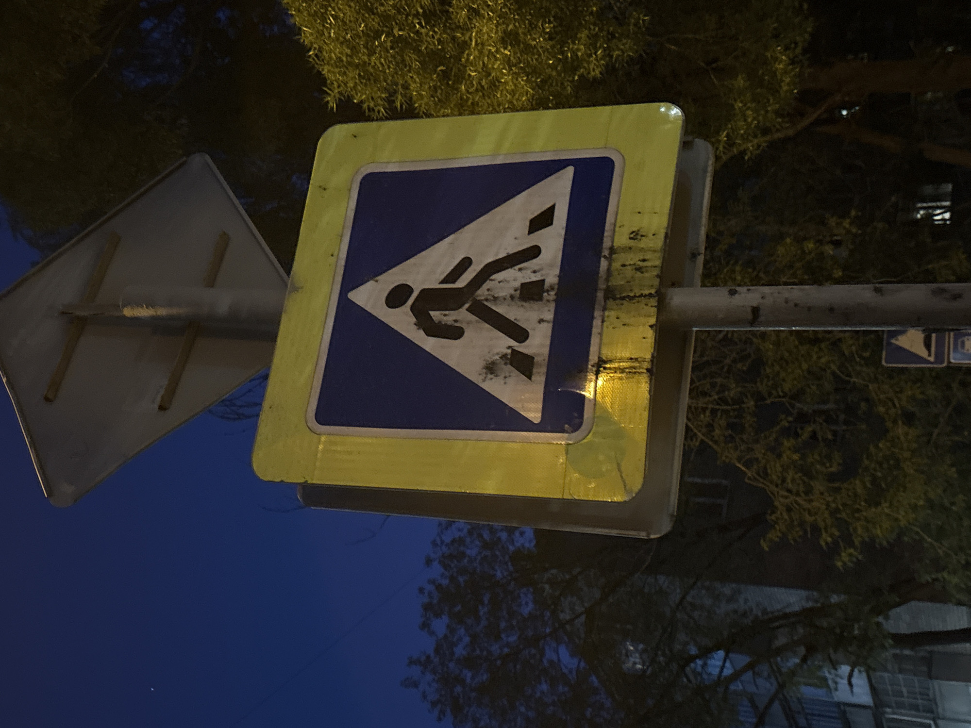 A low-angle, medium shot captures a yellow and blue road sign depicting a stylized pedestrian in mid-stride, within a white triangle, indicating a designated pedestrian crossing. The sign, displaying noticeable wear and imperfections, is affixed to a grey metal pole. To the left, a generic grey road sign is also affixed to the same pole. The backdrop consists of dark blue sky transitioning to darker silhouettes of trees. In the distance, vague architectural features are visible. The location is in Perm, Russia.