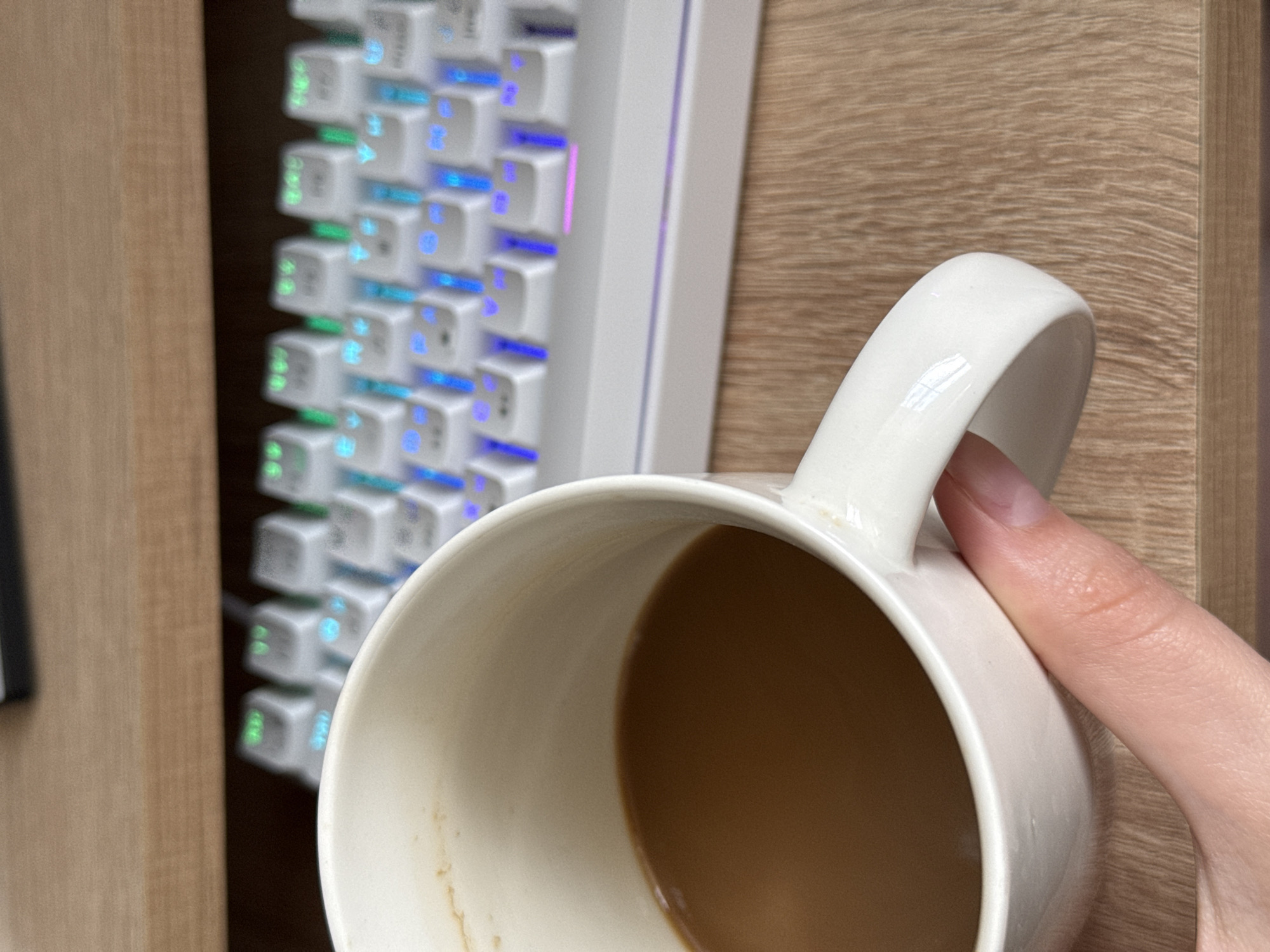 A close-up, eye-level shot shows a white mug filled with coffee, held by a person's hand, positioned in the foreground. The mug is partially obscuring a white mechanical keyboard with RGB backlighting. The keyboard is situated on a wooden surface, possibly a desk. The background is blurred, but the overall impression is that of a workspace. The lighting is natural, likely from a nearby window, creating soft shadows. There are no distinctive landmarks or environmental cues to suggest a specific location in a city.
