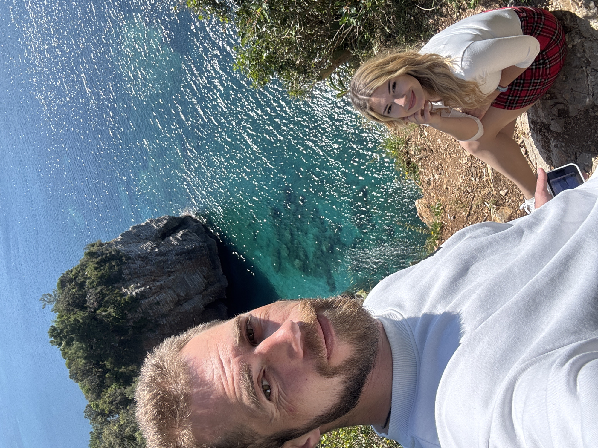 A selfie, taken on a cliff overlooking the Adriatic Sea, features two people. The man, likely holding the camera, is centered in the foreground; he has light brown hair and a beard, and is wearing a white shirt. A young woman with blonde hair is sitting behind him and to the right, wearing a white shirt and a red plaid skirt. She is looking at the camera with a smile, and her hands are posed near her face. The sparkling turquoise sea fills much of the background, and a rocky, tree-covered islet is visible to the left. The vegetation and ground near the edge of the cliff, which is also in the background, appears dry and sparse. The photograph was taken at a viewpoint along the coast in the vicinity of Budva, Montenegro.
