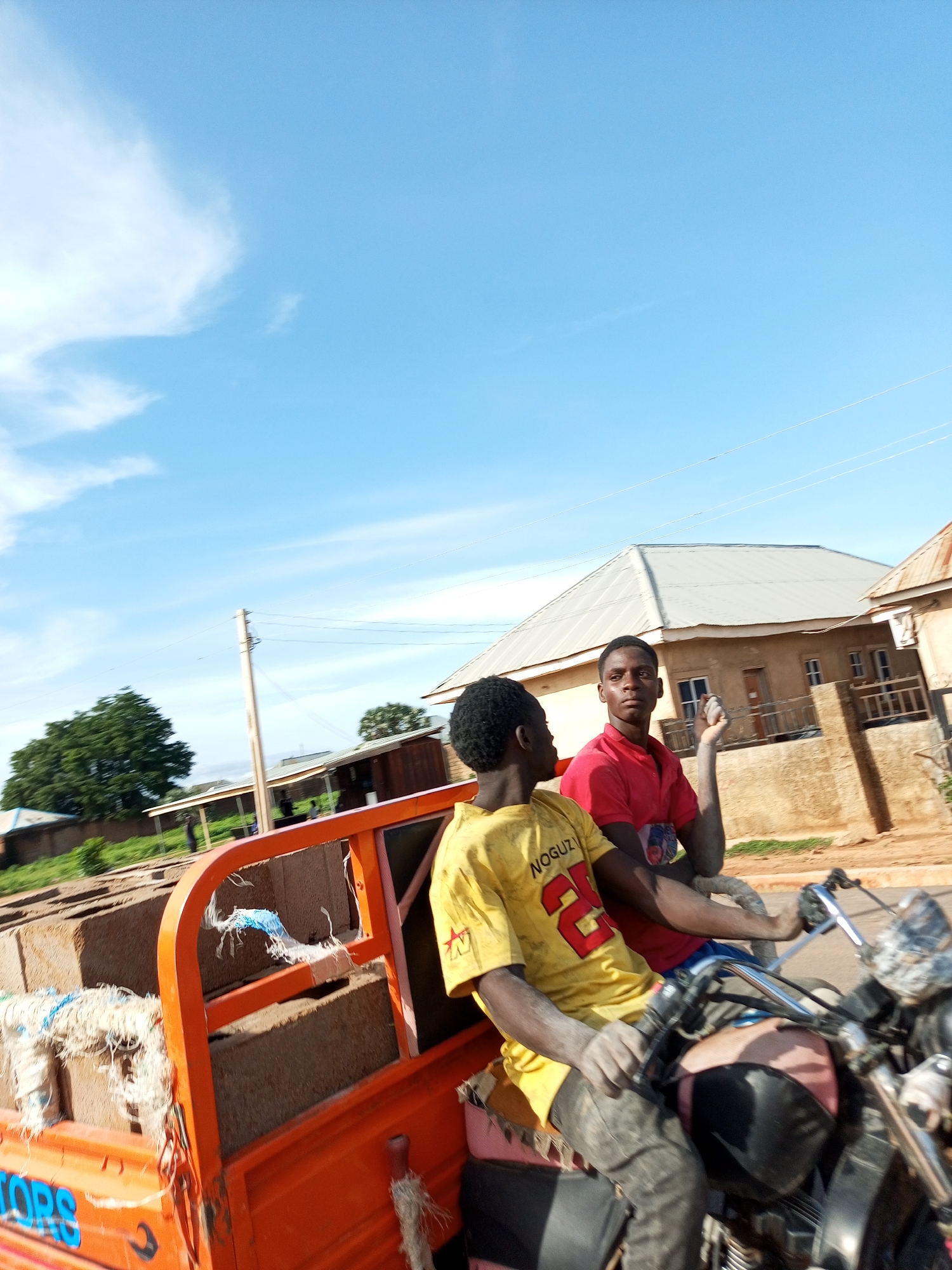 Two young Black men are riding on the back of a three-wheeled orange transport vehicle, possibly a tuk-tuk. The man in the foreground, on the left, wears a yellow t-shirt with red lettering that reads "NOGUZI 2". He also wears dark, dusty pants. The man behind him, on the right, wears a red t-shirt. His left hand is raised and appears dusty. Both men have dark skin and black hair. The vehicle is filled with what appear to be large, rectangular blocks, possibly bricks or building materials, tied down with rope.
The vehicle is moving along a dirt road in a rural or semi-rural area. In the background, there are several buildings with corrugated metal roofs and brown walls. One building has wooden supports. There are also trees and a telephone pole with wires. The sky is bright blue with some scattered white clouds, suggesting it is daytime, likely morning or afternoon, and the weather is clear. The overall impression is one of daily work or transportation in a Nigerian setting. The men appear focused on their journey. There is no clear indication of their emotional state, though their dusty appearance might suggest they are engaged in physical labor. The word "TORS" is partially visible in blue lettering on the side of the orange vehicle.