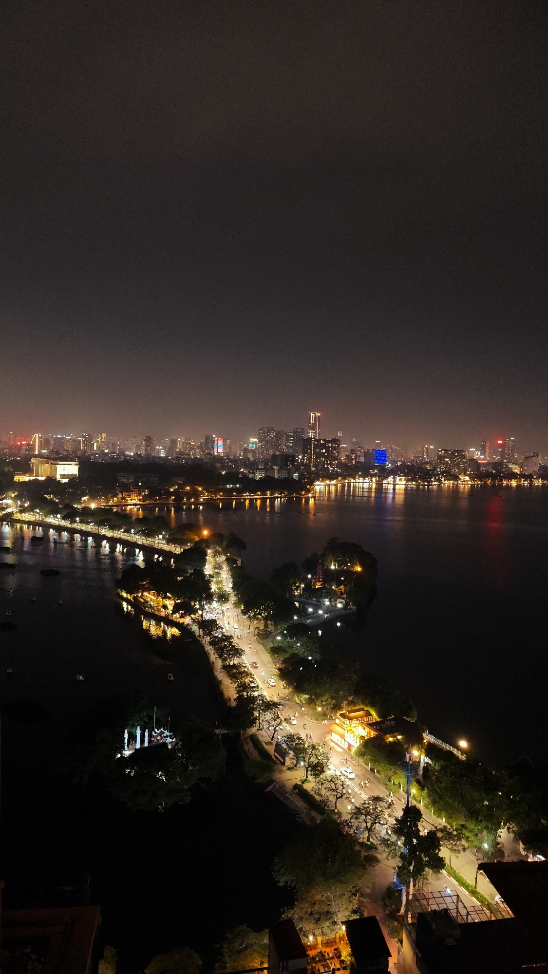 This is a nighttime aerial view of a city skyline and a body of water, likely a lake, with a causeway or bridge extending into it. The city in the background is illuminated by numerous lights from buildings, creating a vibrant and sprawling urban landscape under a dark sky, possibly indicating dusk or night. The water reflects the city lights, creating a shimmering effect.
In the foreground and midground, there are trees lining a road that runs along the edge of the water. The road is lit by streetlights and illuminated by the headlights and taillights of moving vehicles, suggesting a moderate flow of traffic. There are also some structures with illuminated windows and rooftops, possibly restaurants or other buildings. Small boats or watercraft are visible on the water.
Notable details include the time of day, which is clearly night, indicated by the artificial lighting and the dark sky. The weather appears to be clear, as the city lights are sharp and well-defined. The presence of illuminated buildings, water, and a distinct urban layout suggests a significant city location. There are no discernible emotions conveyed in the image, as it is a landscape. No visible text is present in the image. The scene captures the lively atmosphere of a city at night, with the interplay of light and shadow over both land and water. The overall impression is one of urban vitality and the beauty of a city illuminated after dark.