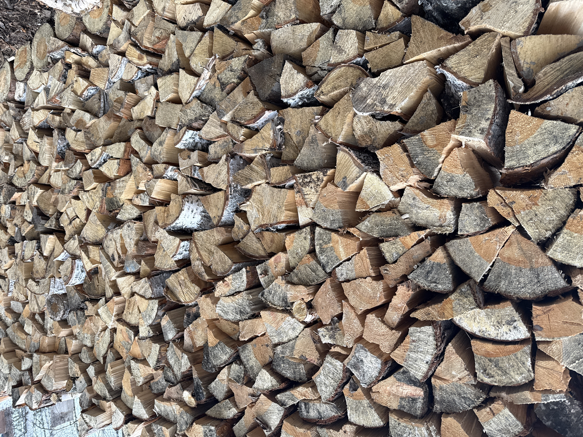 A close-up, high-angle shot shows a large stack of chopped firewood. The firewood consists of irregular pieces of wood, likely birch given the presence of white bark visible on some pieces. The wood pieces are stacked tightly together. The texture of the wood is rough, with visible wood grain and some weathered surfaces. The color of the wood varies from light beige to darker gray and brown tones. The overall impression is that of a large, practical pile of fuel for burning. There is no information about the location in the city of Yekaterinburg, Russia. No people are visible.