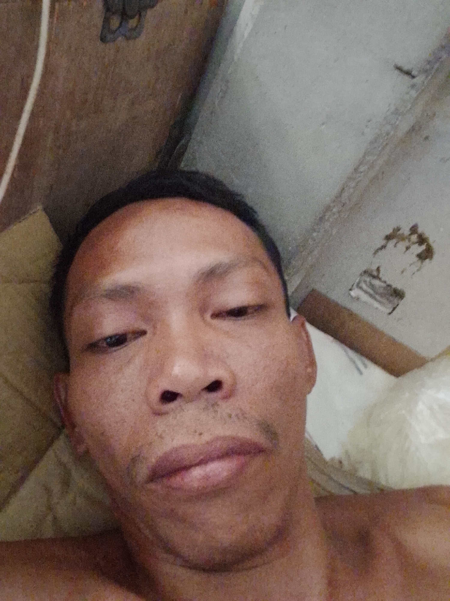 A selfie of a middle-aged Asian male with dark hair and a light brown complexion. He is lying down, with a neutral expression, appearing tired or unwell. The background consists of a weathered wall or ceiling with visible patching and discoloration, and a cardboard box. The location is inferred from the prompt to be Bukit Ketok, Indonesia.