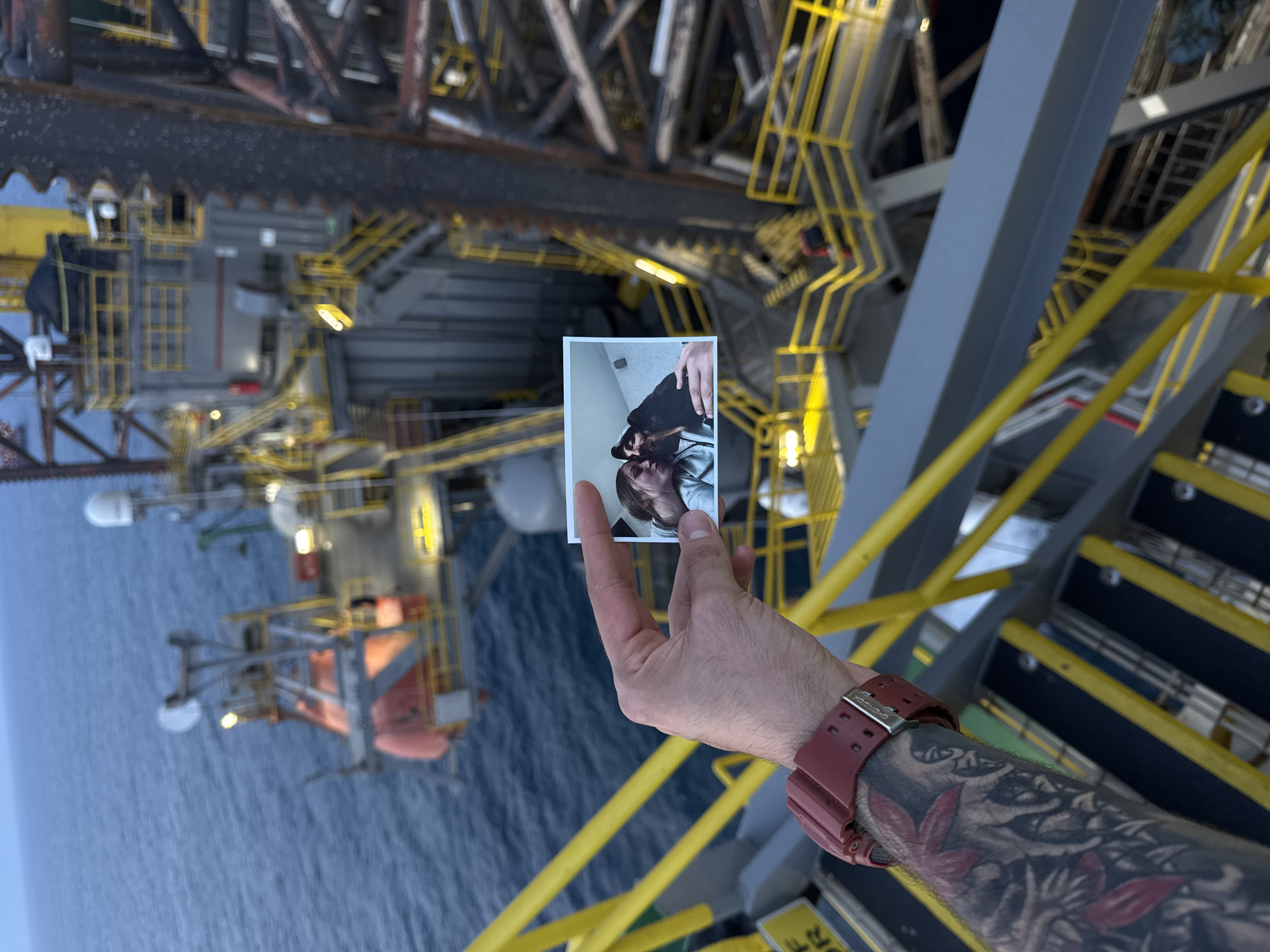 An individual's hand, adorned with tattoos and a red watch, holds a small photograph in front of an offshore oil rig. The photograph displays a close-up image of two individuals, a man and a woman, appearing to be a couple. The man in the photo has a beard and is wearing a dark cap and a light-colored hoodie. The woman, also in a light-colored hoodie, is leaning her head towards him. The background of the main image shows the vast expanse of the ocean and a portion of the oil rig, with its complex structure of metal platforms, stairs, and railings, bathed in the soft light of dusk. The location is not specified as being in the city of Unknown, Italy.