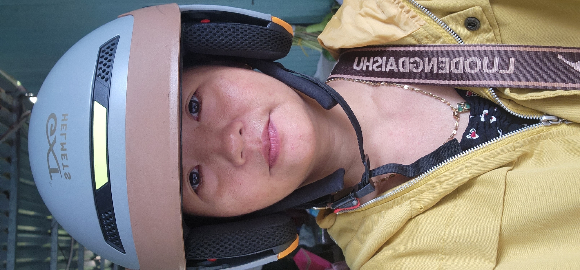 A medium shot image shows a woman from the chest up, wearing a light blue and tan helmet with black ear pads. She is also wearing a yellow hooded jacket zipped up, with a black shirt underneath. A gold necklace with a green pendant is visible. A patterned strap with text in a foreign language crosses her chest. The background appears to be outdoors, with a corrugated metal roof and what looks like a wall to the right. The lighting suggests it is daytime.