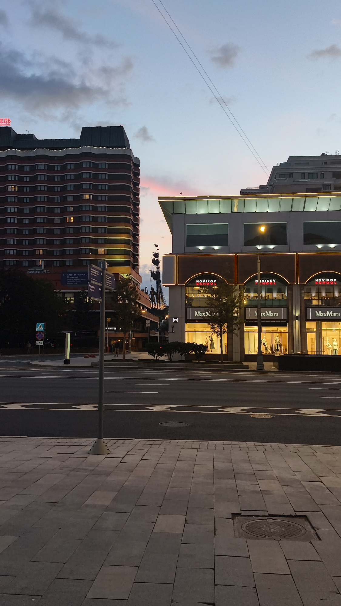 This image depicts a street scene in Moscow, Russia, likely in the evening given the dusky sky and illuminated buildings. On the left, a tall, multi-story brick building with horizontal bands of windows is visible. Its top section features a darker hue and decorative lighting along its edge. To the right, a modern building with a white facade is illuminated by warm interior lights, highlighting arched storefronts. These storefronts display signage for "NOVIKOV SCHOOL" and "MaxMara". The street in the foreground is paved, and a sidewalk with similar paving stones is to the left of the road. Traffic lights and street signs are present, including one with Cyrillic text. A pedestrian crossing is marked on the road. The sky is a gradient of pale blue and grey, with hints of pink from a setting sun or ambient light. Power lines stretch across the sky. The scene is devoid of prominent human activity, suggesting a relatively quiet moment in the city.
