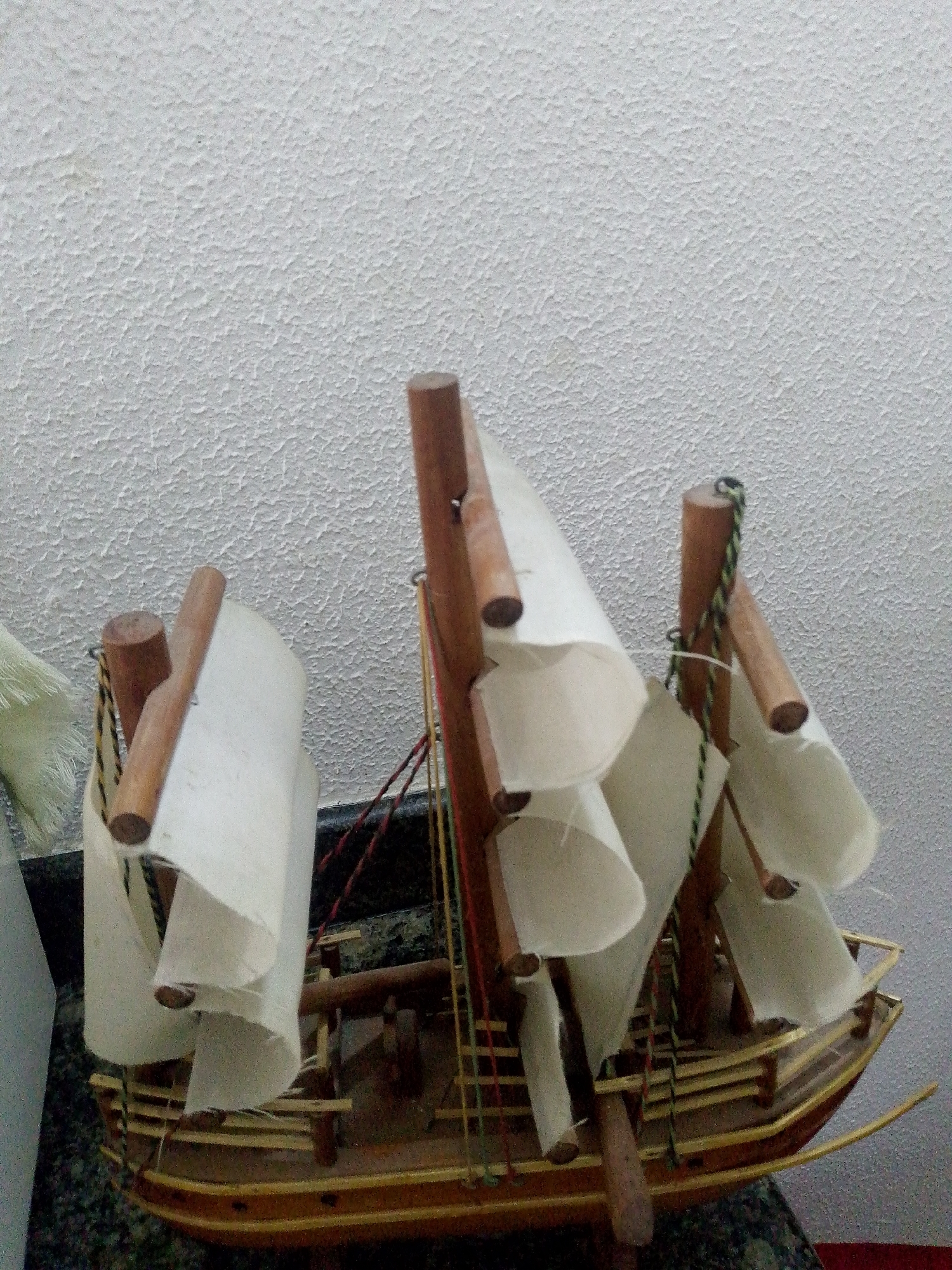 A miniature wooden sailing ship with white paper sails sits in front of a textured white wall. The ship has three masts with wooden pegs at the top and bottom. Multiple colored strings connect the masts and hull. The ship rests on a dark, patterned surface. The location is unknown.
