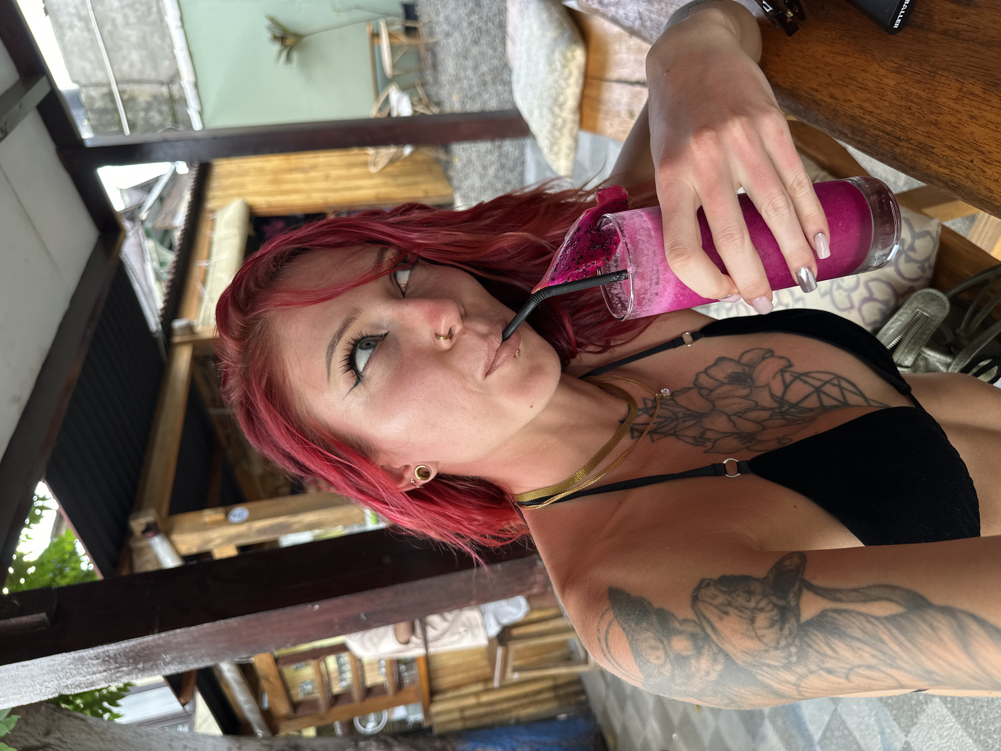 A person with red hair and visible tattoos is drinking a pink beverage with a straw from a glass. The person has facial piercings and is wearing a black top and gold necklaces. The background suggests an outdoor or semi-outdoor location with wooden structures, possibly a cafe or restaurant. The precise city in Indonesia is indeterminate from the image.