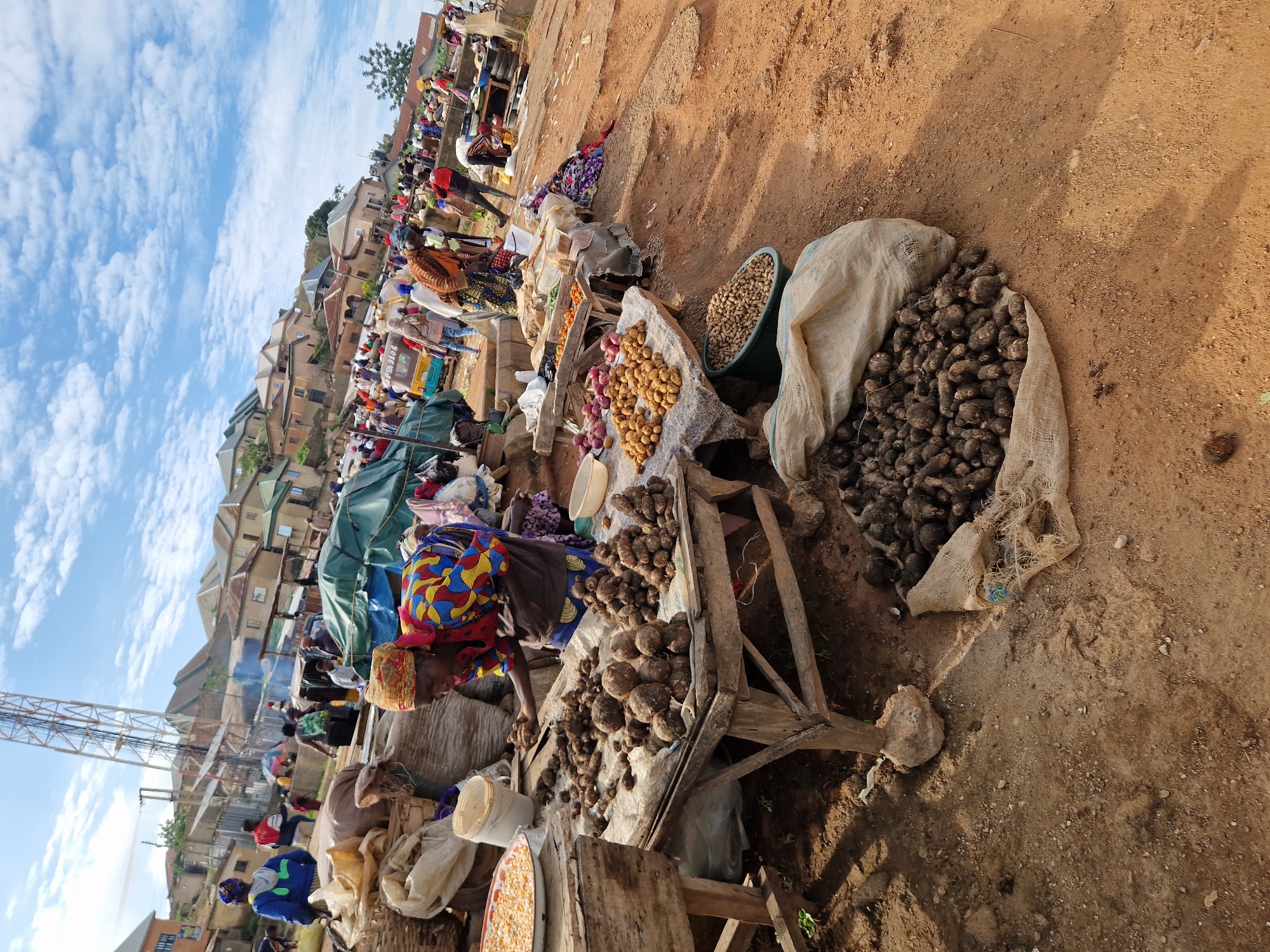 In Kabong, Nigeria, a bustling outdoor market is captured under a partly cloudy sky. The scene is dominated by a woman in a vibrantly patterned blue, yellow, red, and black outfit, bent over a wooden stall laden with dark, irregularly shaped root vegetables. She appears to be selecting or arranging them. The stall also displays a round tray of corn and a white bucket. To her left, another stall features a pile of potatoes and red onions. In the foreground, a large burlap sack spills over with similar root vegetables, resting on reddish-brown dirt ground. Behind the stalls, a diverse crowd navigates the market, with some individuals carrying goods or interacting. A tall communication tower is visible in the background, juxtaposed with a cluster of houses and buildings that form a hillside. A small blue and yellow motorized tricycle is parked amongst the shoppers. The environment suggests a warm, possibly dry, climate.