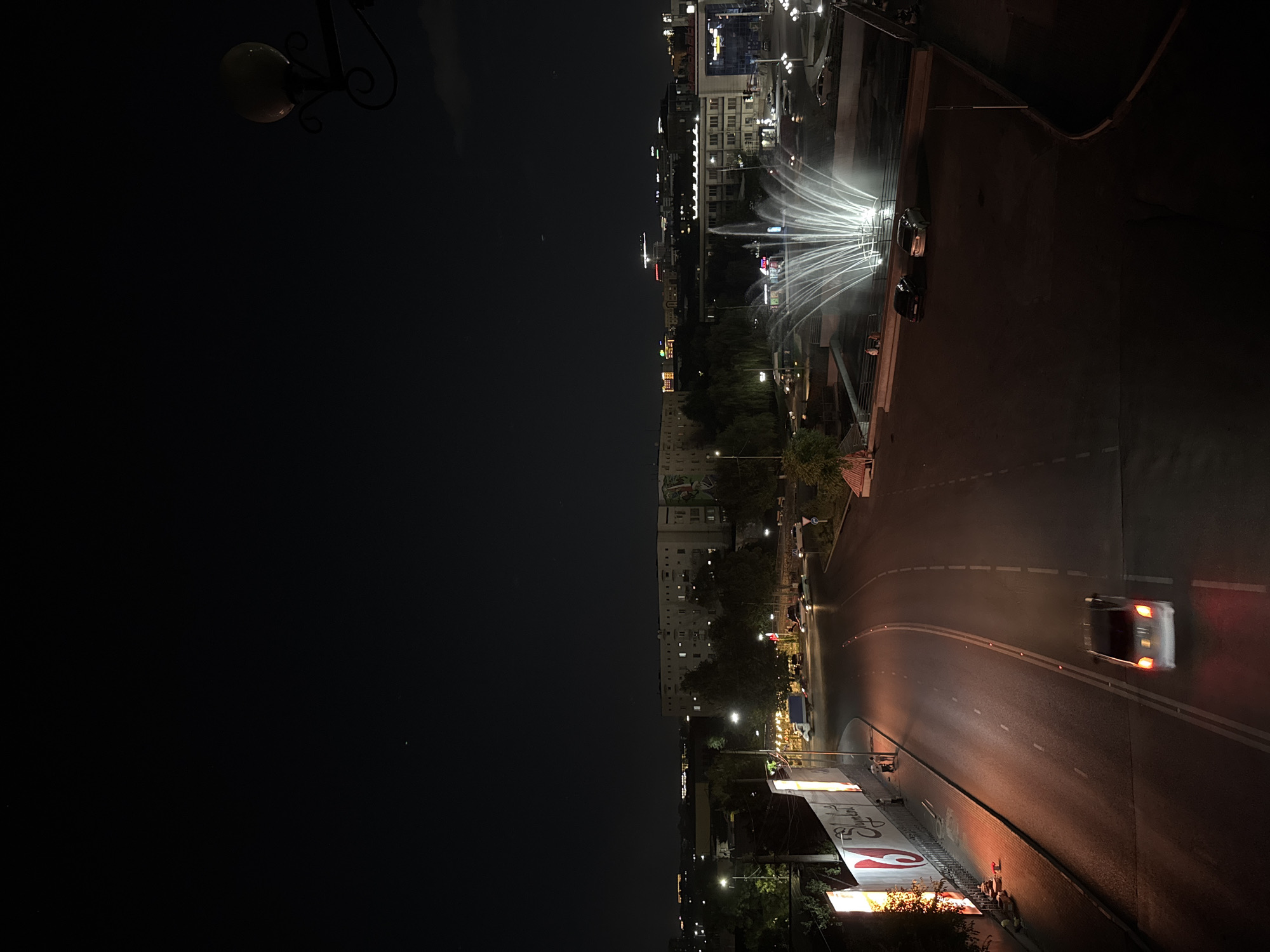 The image captures a nighttime view of a street in Shymkent, Kazakhstan. A highway with illuminated lanes curves through the frame, with a single white car in motion on the road. To the left of the highway, a tall, illuminated billboard displays advertisements. Further into the scene, a large fountain with brightly lit water jets is active in a plaza. Several cars are parked near the fountain, and a few individuals can be seen walking nearby. In the background, residential and commercial buildings are visible, illuminated by streetlights and interior lighting, with a dark, starless sky above. A decorative streetlamp with an ornate metal base is visible in the upper right corner.