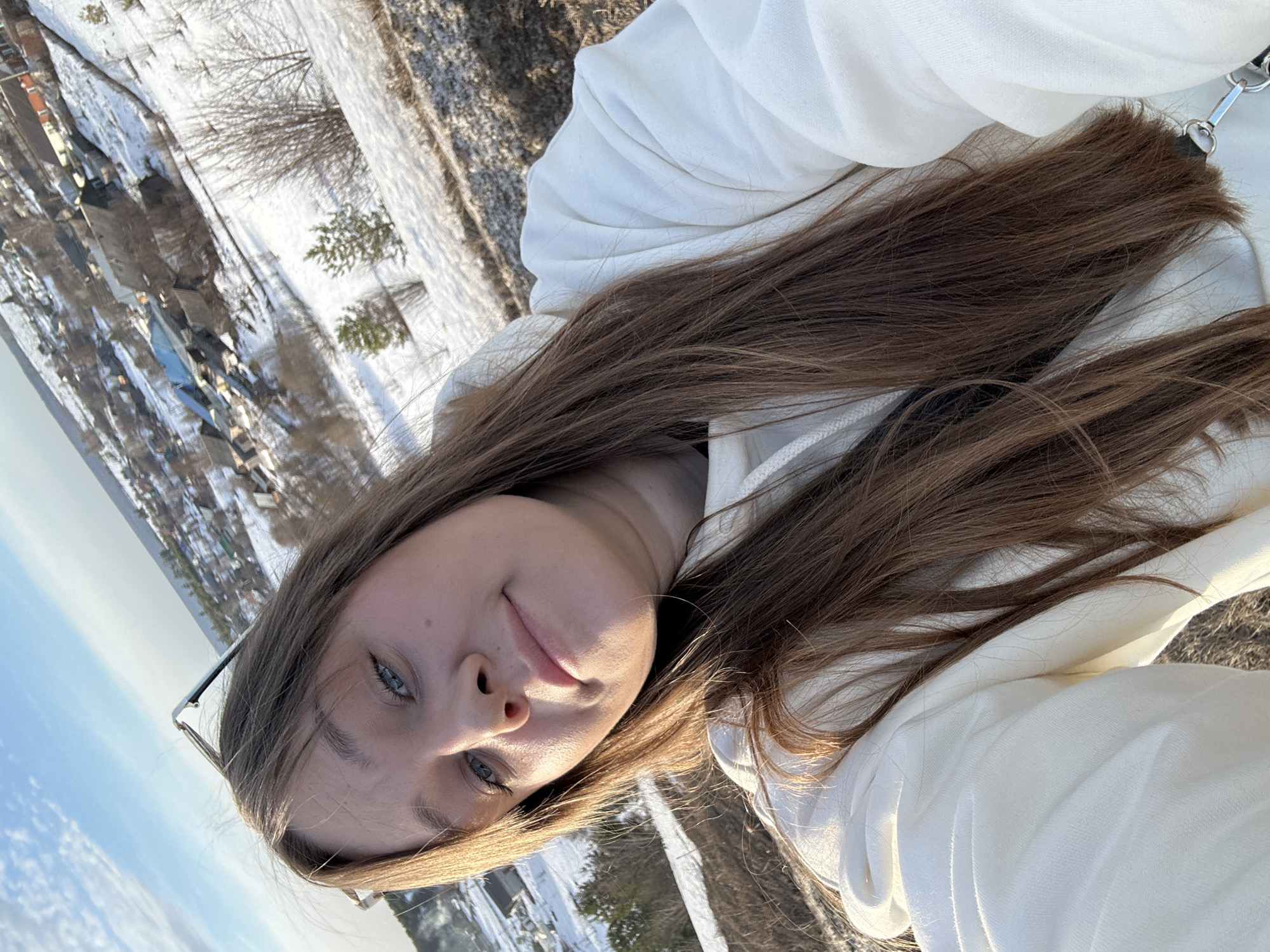 A female subject is depicted in a close-up selfie photograph, taken outdoors in the city of Фоки, Russia. She has long, straight brown hair, blue eyes, and a fair complexion, and is looking directly at the camera with a subtle smile. Sunglasses are perched on top of her head, and she is wearing a white hooded sweatshirt. A dark strap is visible across her left shoulder. The background reveals a snow-covered landscape from an elevated perspective, showing a village nestled on a gentle slope below. The environment includes sparse dry vegetation and bare trees in the foreground and midground, with houses of varying roof colors (dark grey, blue, red) populating the village. The sky above is partly cloudy blue, and the scene is illuminated by natural sunlight, casting a warm glow on the subject's right side and parts of the snowy terrain.