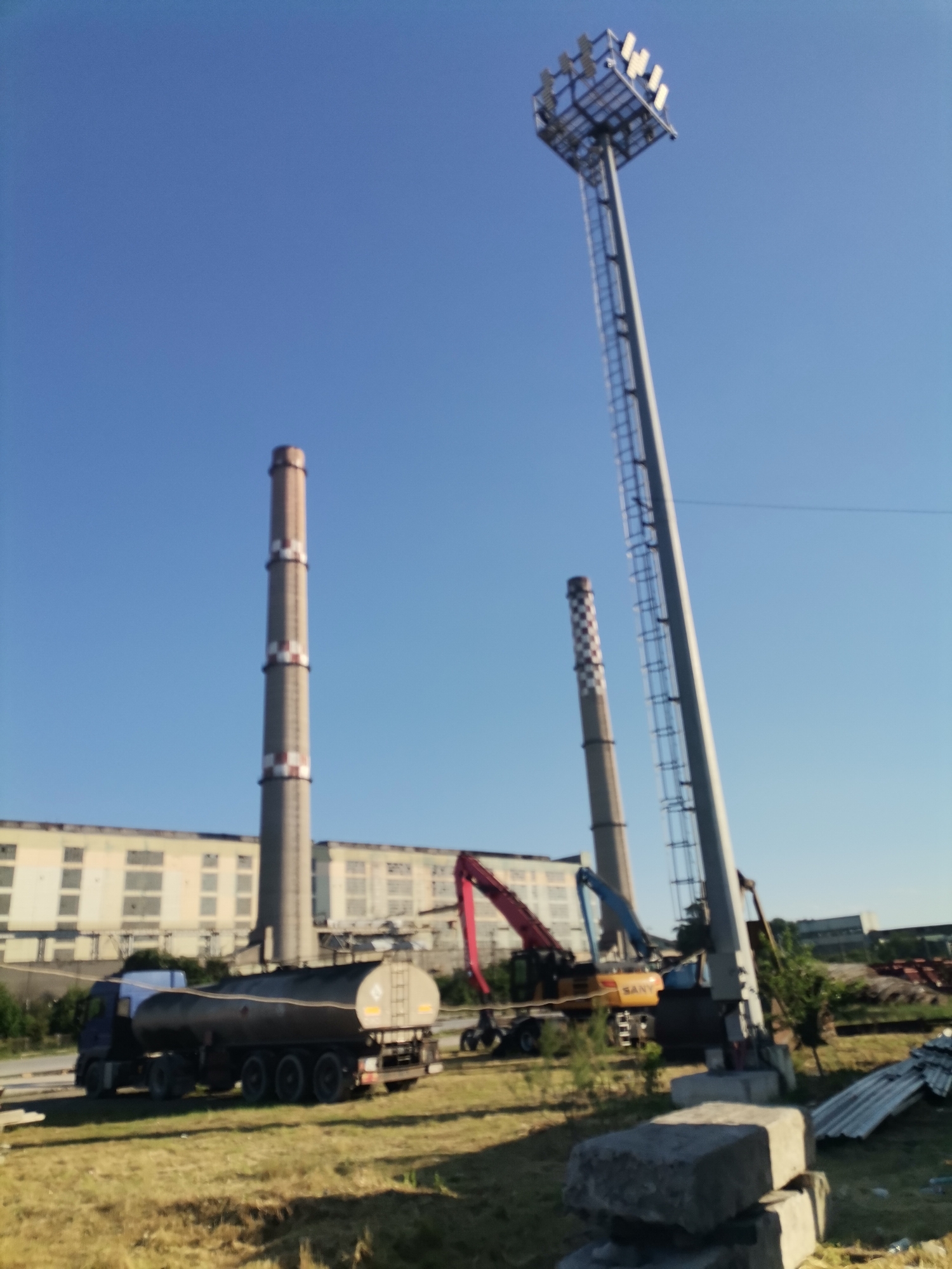 The image depicts an industrial area in Ezerovo, Bulgaria, with two smokestacks towering over a large, low-rise building. A fuel tanker truck is parked in the foreground. Adjacent to the truck is construction equipment, including a yellow crane with a red arm and a blue arm. A tall light pole with a ladder and a set of lights at the top stands beside the crane. In the immediate foreground, concrete blocks and corrugated metal sheets are scattered on the ground. The scene is set under a clear blue sky, suggesting a sunny day.