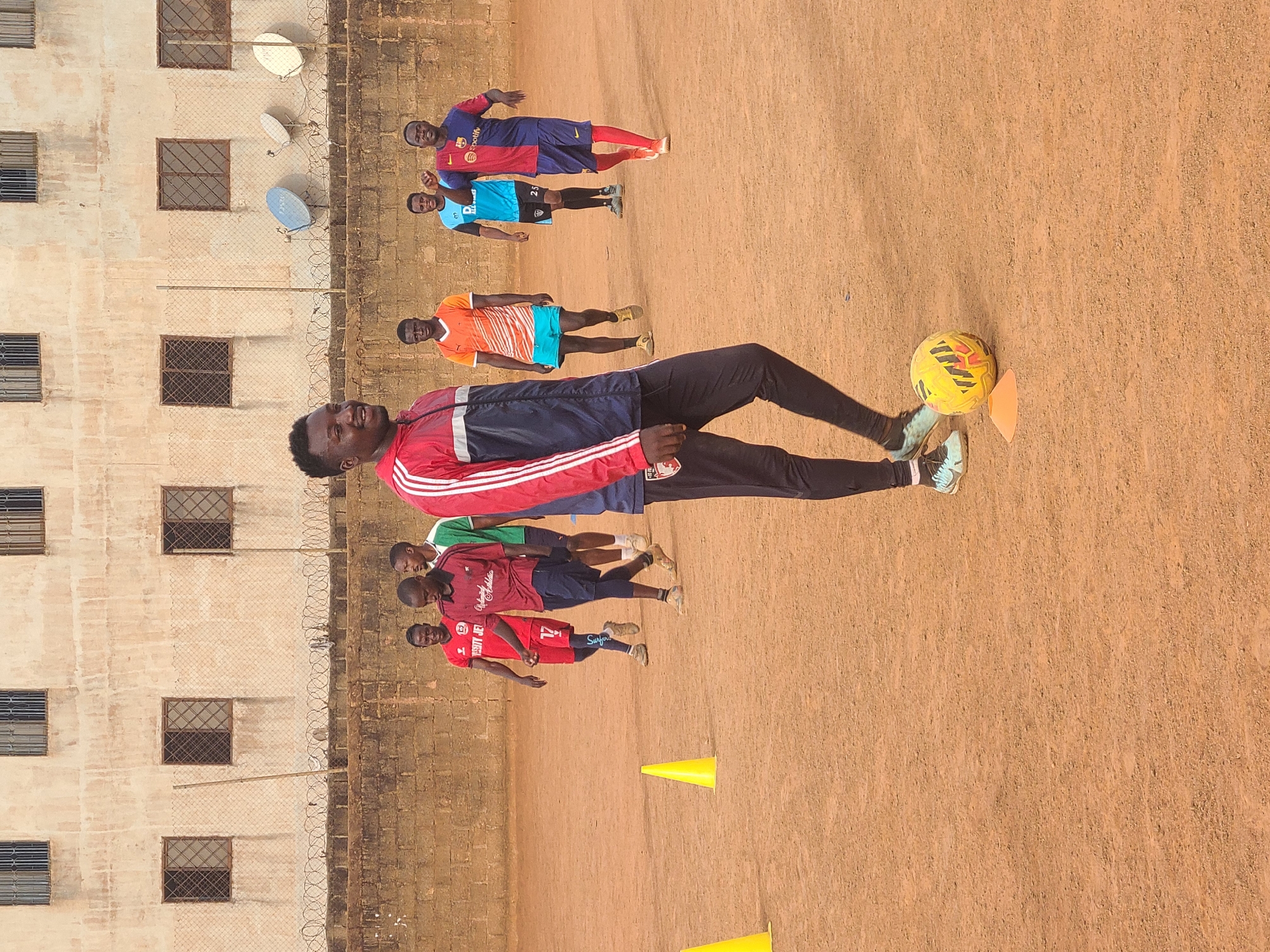 A group of men and boys are engaged in a soccer practice session on a dirt field in Barakin Lerawi, Nigeria. One man is in the foreground, facing the viewer, wearing black pants and a red, white, and navy-blue jacket. He is standing near a yellow soccer ball and an orange cone. Behind him, four men are in red shirts. Three men in the background are wearing a Barcelona FC jersey, an orange jersey, and a blue jersey. In the background is a building with a wire fence along the top. Three satellite dishes are mounted on the building. The ground is a light reddish-brown dirt. The sky is bright.