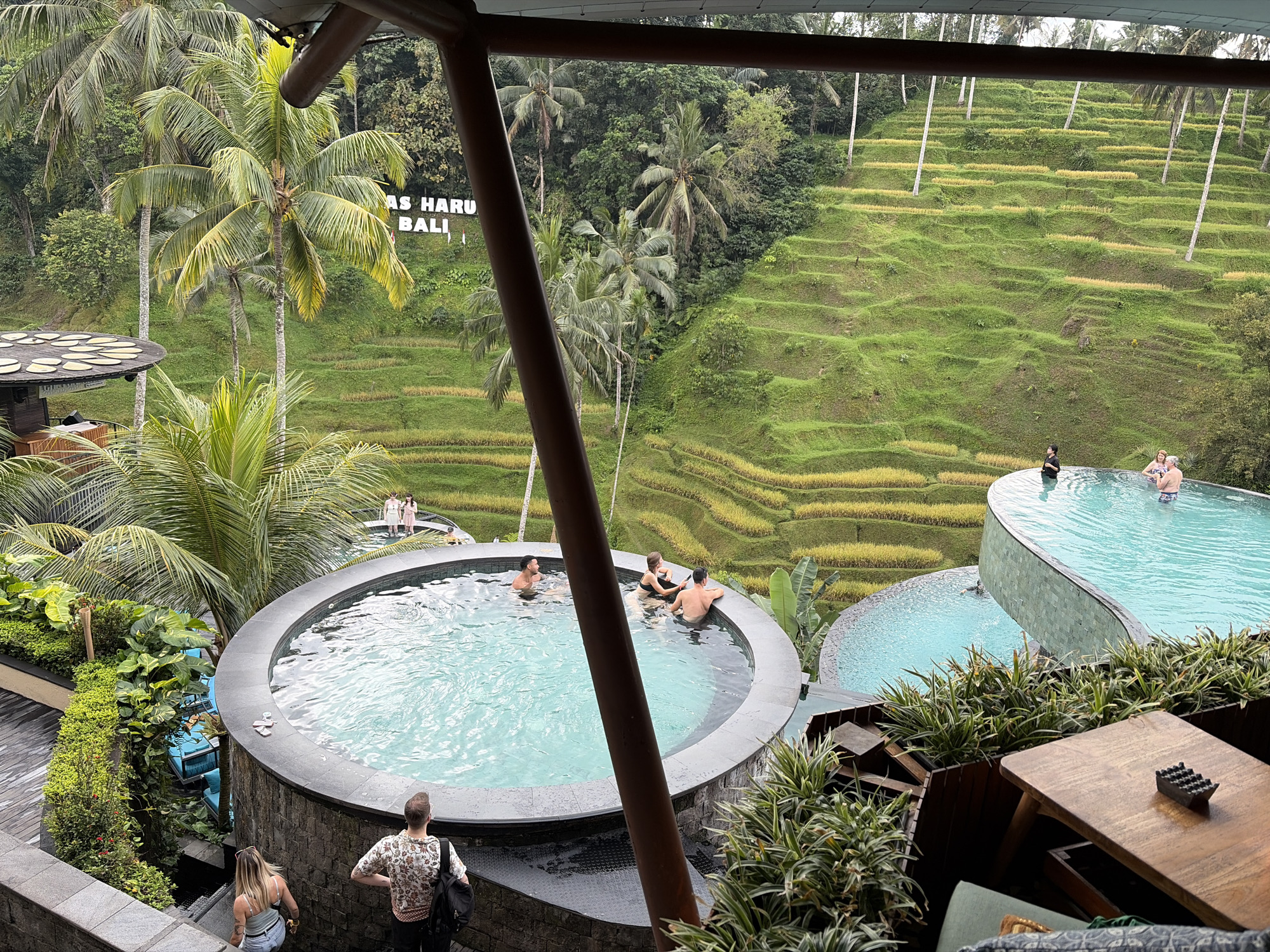 The image depicts a scenic overlook in Tegallalang, Indonesia, showcasing lush green rice terraces cascading down a hillside. In the foreground, a circular infinity pool with crystal-clear blue water is visible, with several people casually interacting within it. A man and a woman are standing on a stone staircase to the left, descending towards the pool area. Further back, an irregularly shaped infinity pool overlooks the rice terraces, with a few individuals relaxing in the water. A wooden table with an ashtray sits on a deck to the right, surrounded by dense green foliage. In the background, the iconic "NAS HARU BALI" sign is partially visible amidst palm trees and dense jungle vegetation. The overall atmosphere is one of relaxation and natural beauty, with the geometric patterns of the rice terraces contrasting with the organic shapes of the surrounding landscape.