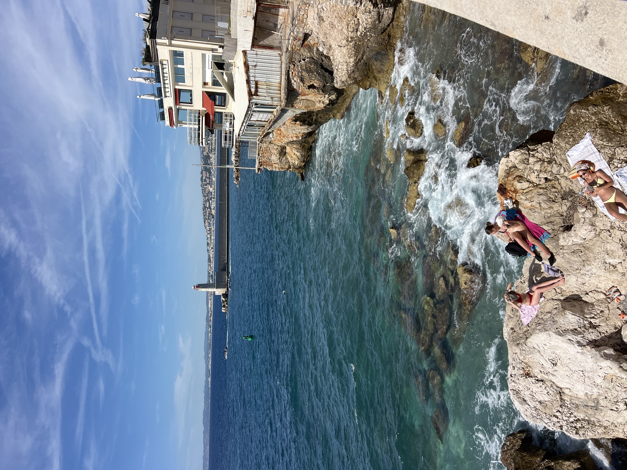 This is a photograph taken on a sunny day in Nice, France, likely from a coastal path overlooking the Mediterranean Sea. The scene captures a section of the coastline with a building perched on a rocky outcrop. In the foreground, several people are sunbathing on large rocks by the water's edge. One person in a red bikini is seated, adjusting their hair, while another person in a yellow bikini is lying down, holding a phone. A third person in a bikini is partially visible to the left, sitting with a towel. The turquoise water crashes against the rocks, creating white foam. Further out in the sea, a lighthouse stands on a pier, with a distant coastline visible on the horizon. The building on the cliff has multiple levels, balconies, and awnings, suggesting it is a restaurant or hotel. The sky is blue with scattered white clouds. The overall atmosphere is one of leisure and enjoyment of the seaside.