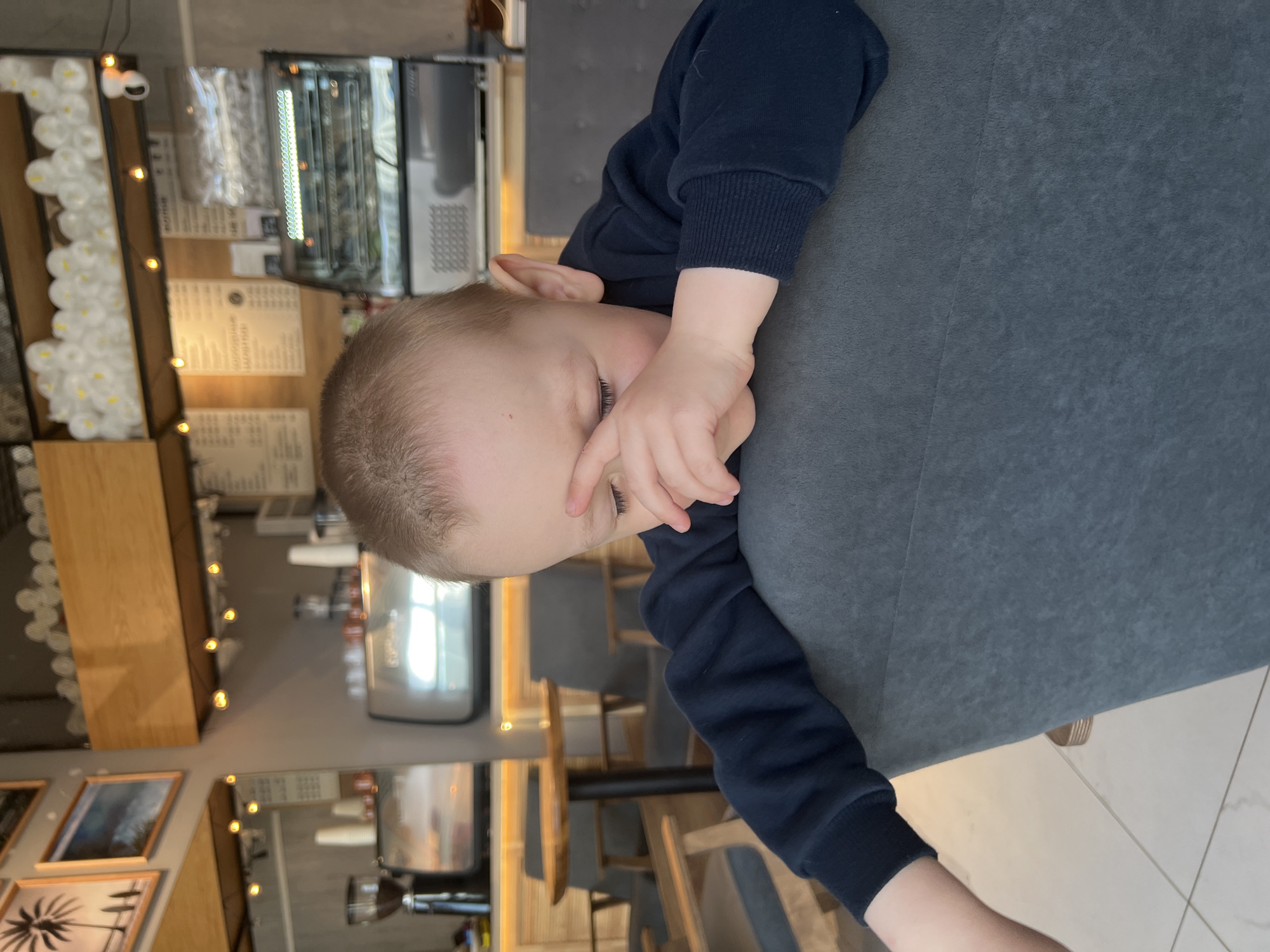 A young child, likely a toddler, is lying with their head on a gray chair in what appears to be a café or restaurant. The child is wearing a dark blue, long-sleeved shirt. The child has fair hair and is partially covering their face with one hand.
The background reveals the interior of the establishment. A wooden shelf displays various items illuminated by string lights. A menu board hangs on the wall. Display cases with contents visible can be seen. Tables and chairs are arranged in the dining area.
Based on the presence of the menu and design details, the location is likely a café or restaurant in the city of Mytishchi, Russia.