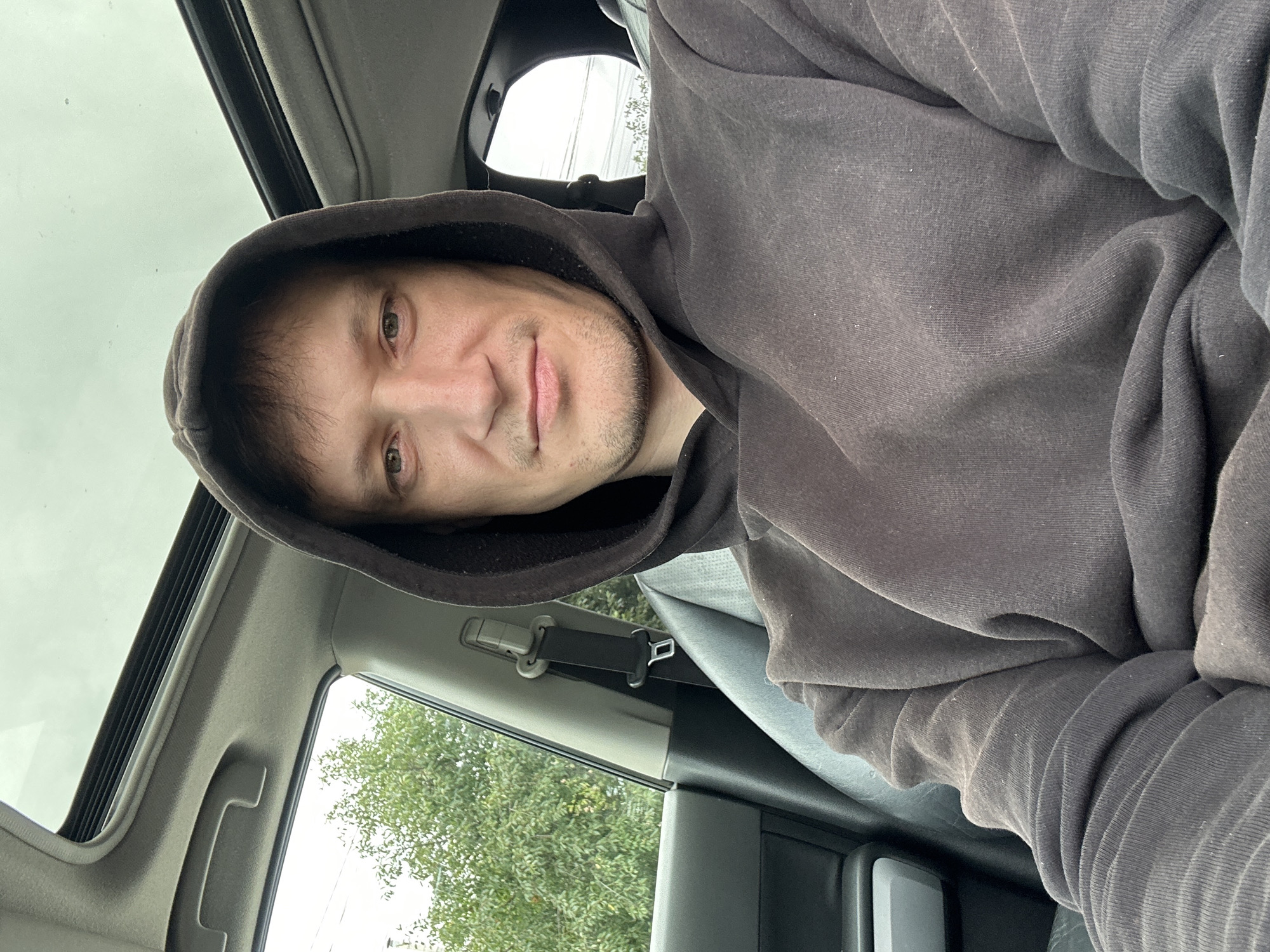 The image shows a selfie taken from inside a car. A man wearing a dark grey hoodie is seated in the driver's seat. The sunroof of the car is open, allowing natural light to illuminate the scene. Outside the car, through the open sunroof and side windows, green trees and a cloudy sky are visible. The interior of the car includes a black leather seat and a grey seatbelt. The location is identified as Makiv, Ukraine.