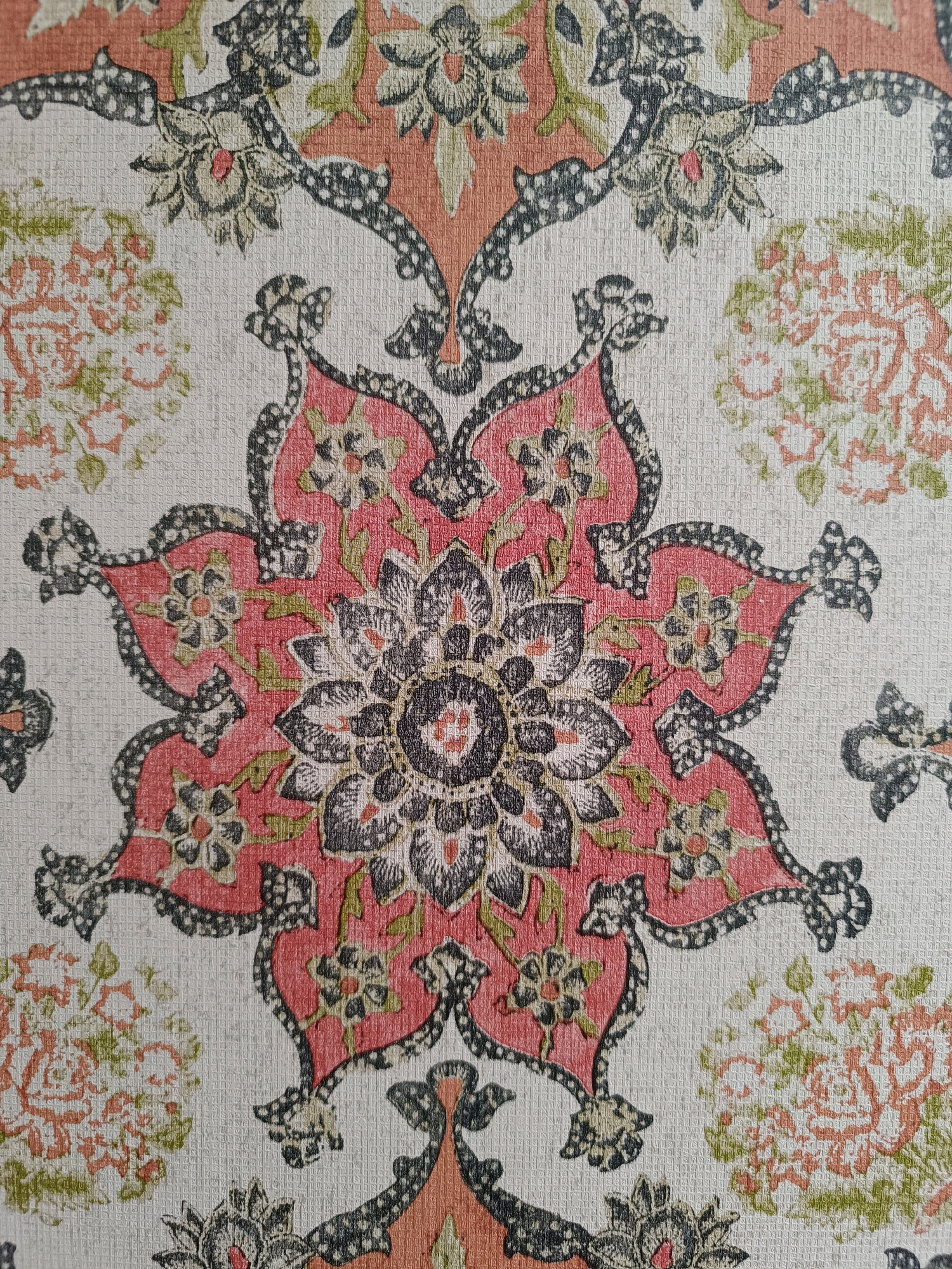 This is a close-up, high-angle, full-frame photograph of a textured fabric with a repeating floral pattern. The dominant colors are shades of coral pink, muted green, black, and a light cream or beige background. The pattern features a large, central eight-pointed floral motif in coral and black, surrounded by smaller floral elements and swirling, paisley-like shapes in black and green. Scattered throughout the background are clusters of smaller, less defined floral shapes in coral and green. The texture of the fabric is visible, appearing to be a woven material with a slightly raised surface, possibly linen or a textured cotton blend. The lighting is even and appears to be natural, with no harsh shadows. The image is purely a detailed study of the textile design and texture, with no indication of any specific location or context, including the city of Srinagar, India.