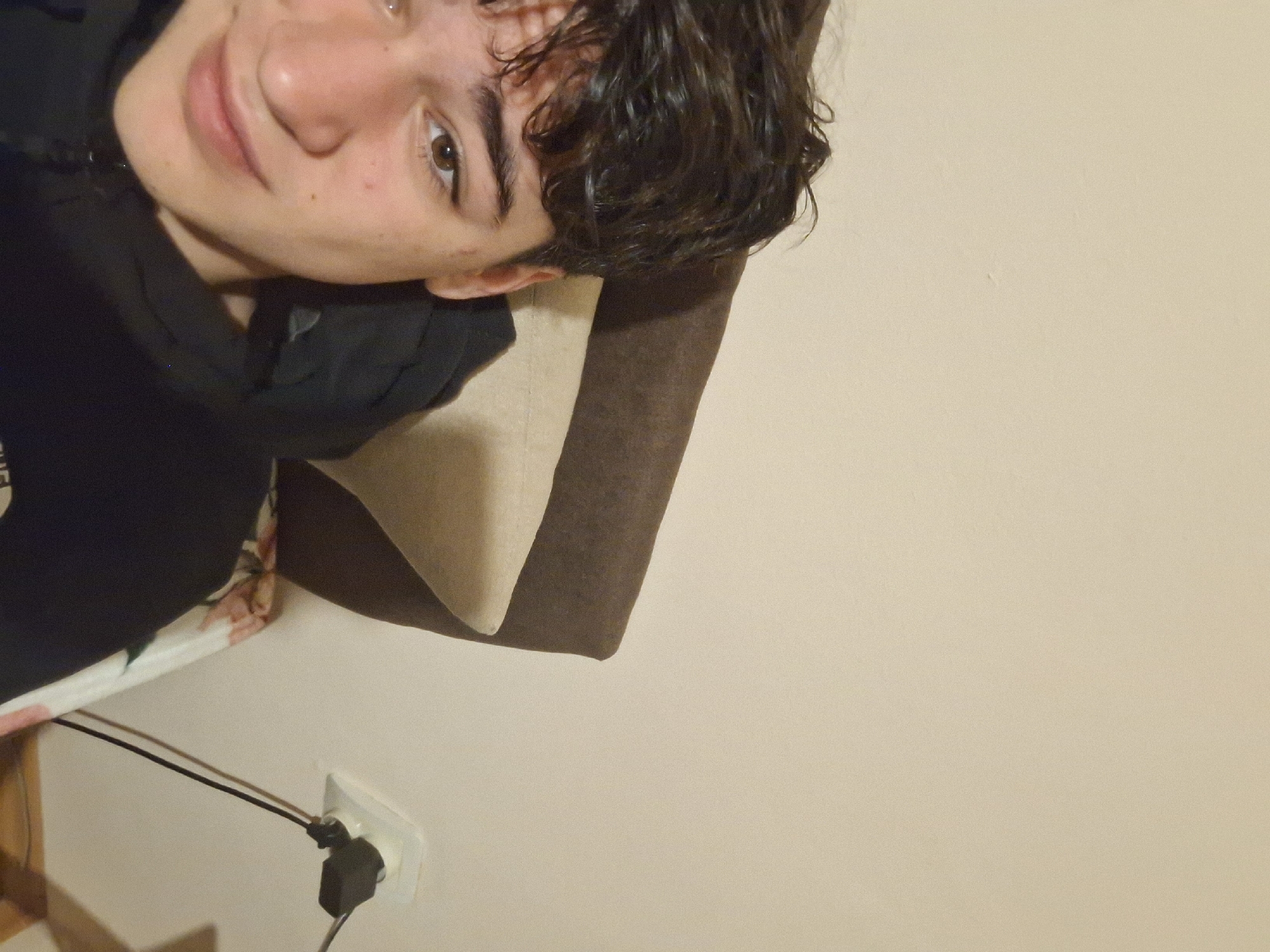A young person with short, dark, curly hair is angled towards the camera, appearing to rest their head against a pillow with a brown and tan cover. They are wearing a dark-colored garment, possibly a jacket or hoodie. To the lower left of the frame, a white electrical outlet is visible with a black plug inserted. The background is a plain beige wall. There is no information available to definitively place this scene in Burgas, Bulgaria.