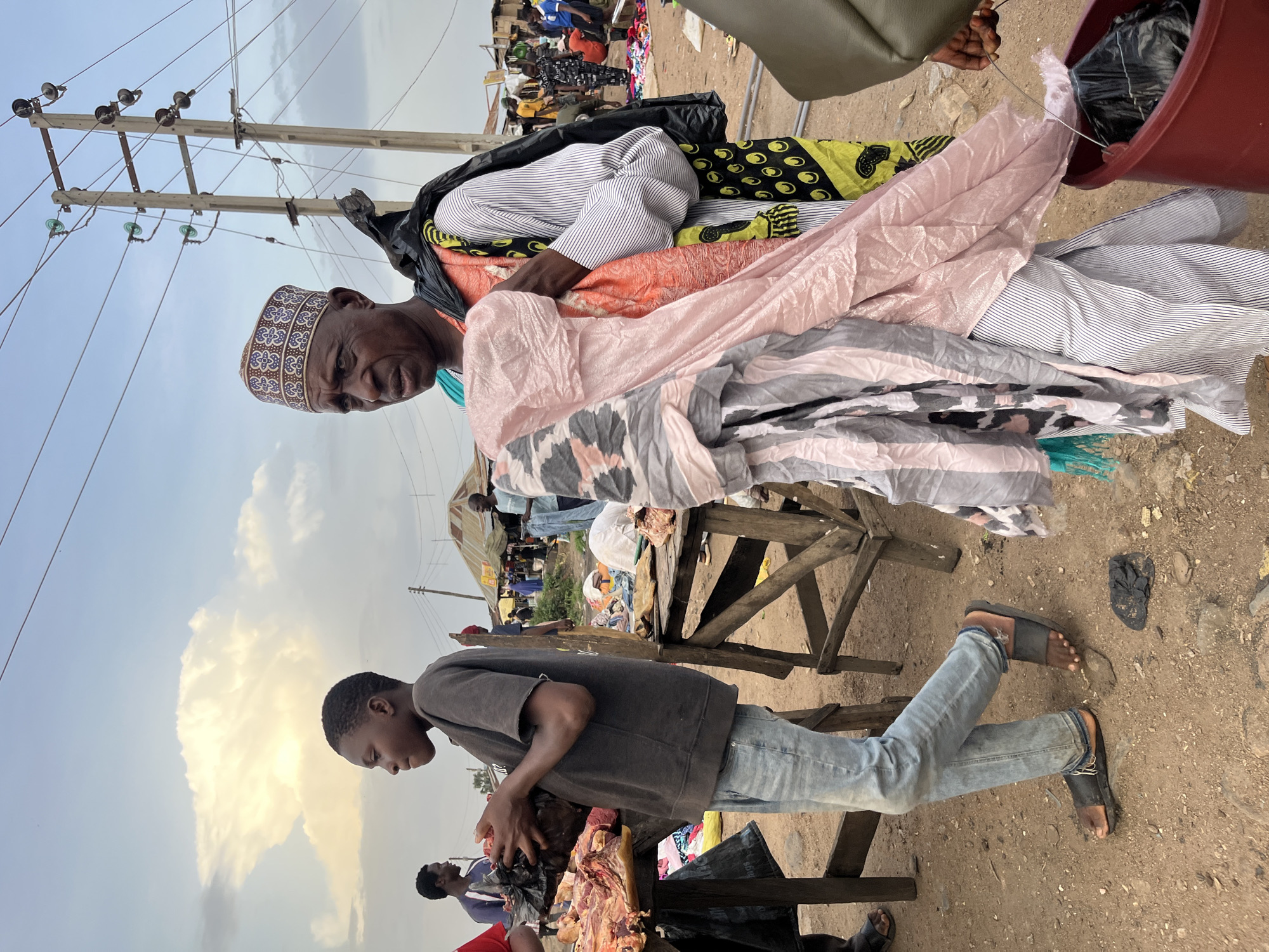 In Bukuru, Nigeria, a marketplace scene unfolds under a cloudy sky. A young boy, clad in a dark t-shirt and light blue jeans, holds a black plastic bag filled with raw meat, standing beside a wooden stall laden with more meat. To his right, an older man wearing a patterned cap and a striped shirt carries a black trash bag slung over his shoulder and draped with colorful fabrics, including a bright orange and a pink garment. He is also seen holding a bundle of fabric. In the background, other vendors and shoppers are visible amidst market stalls and overhead electrical wires. The ground is dusty earth, and scattered debris is present.