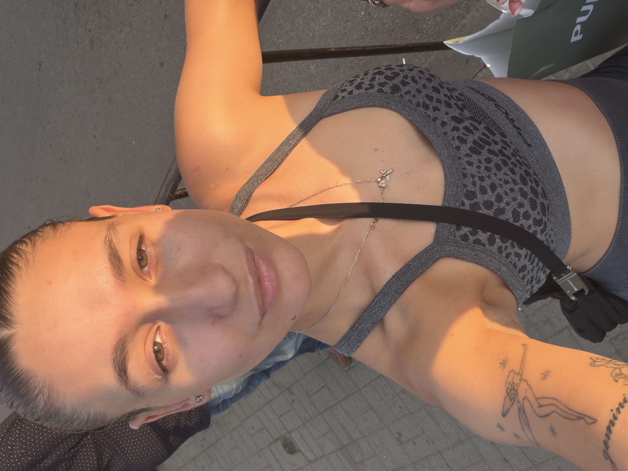A young woman with her hair pulled back in a bun, wearing a grey leopard-print sports bra, poses for a selfie. The sunlight casts a warm glow on her face and arm. She has a tattoo on her upper left arm. Behind her, a person in a dark patterned shirt and jeans is visible, along with a grey paved ground and a metal railing. The scene appears to be outdoors, possibly in a public area of Sofia, Bulgaria. A green shopping bag with white lettering is partially visible in the lower right corner.
