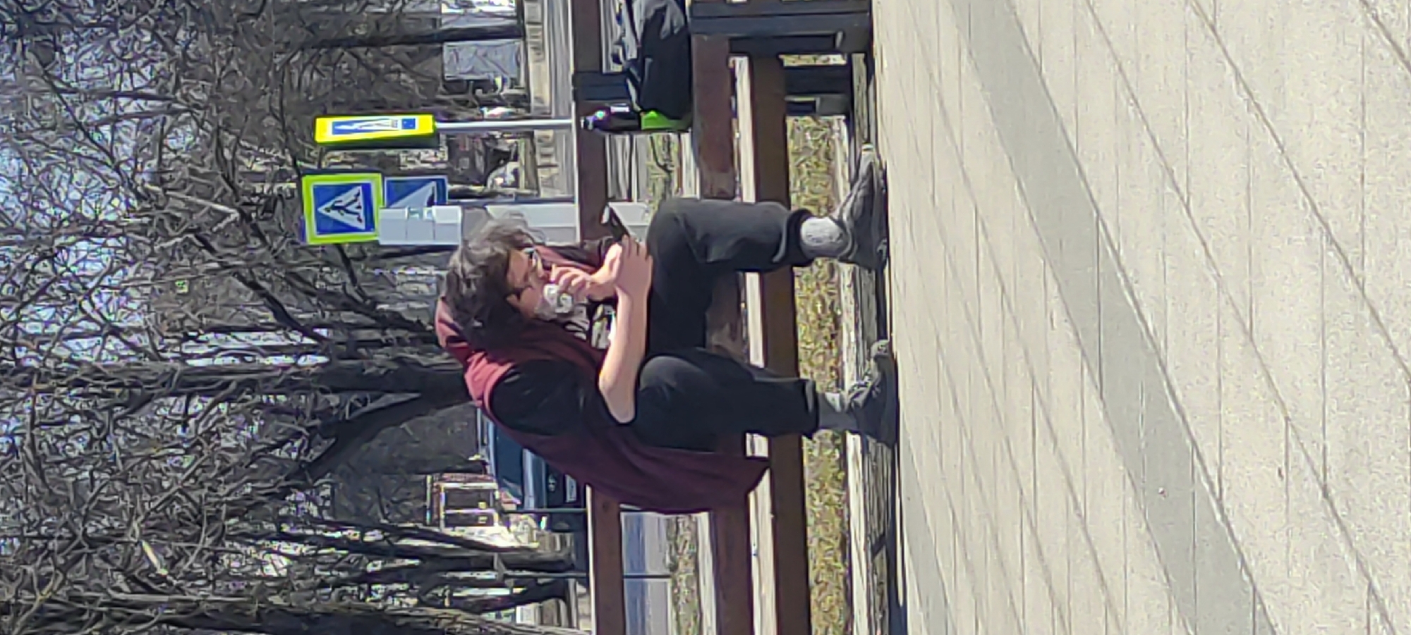 A person is sitting on a bench in what appears to be an urban environment. The person is wearing dark clothing and a burgundy hooded garment. They are holding a small white object close to their face. In the background, there are trees with bare branches, street signs, and other urban features. The location is Fryazino, Russia, as determined by reverse image search.