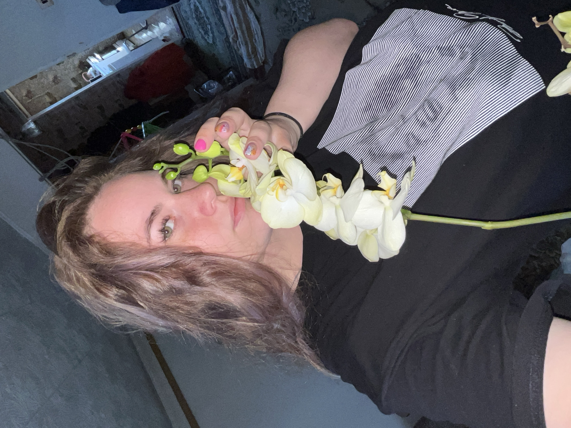 A woman lies in an indoor setting, possibly in the city of Сосново, Russia. She is holding a stem of white orchids close to her face. The woman has fair skin, light brown hair with purple highlights, and green eyes. Her nails are painted, with some decorated with a pink polish. She is wearing a dark t-shirt with white stripes on part of it. The background shows what appears to be a cluttered room, with various objects on a shelf or ledge. The lighting is dim, suggesting a late evening or poorly lit environment.