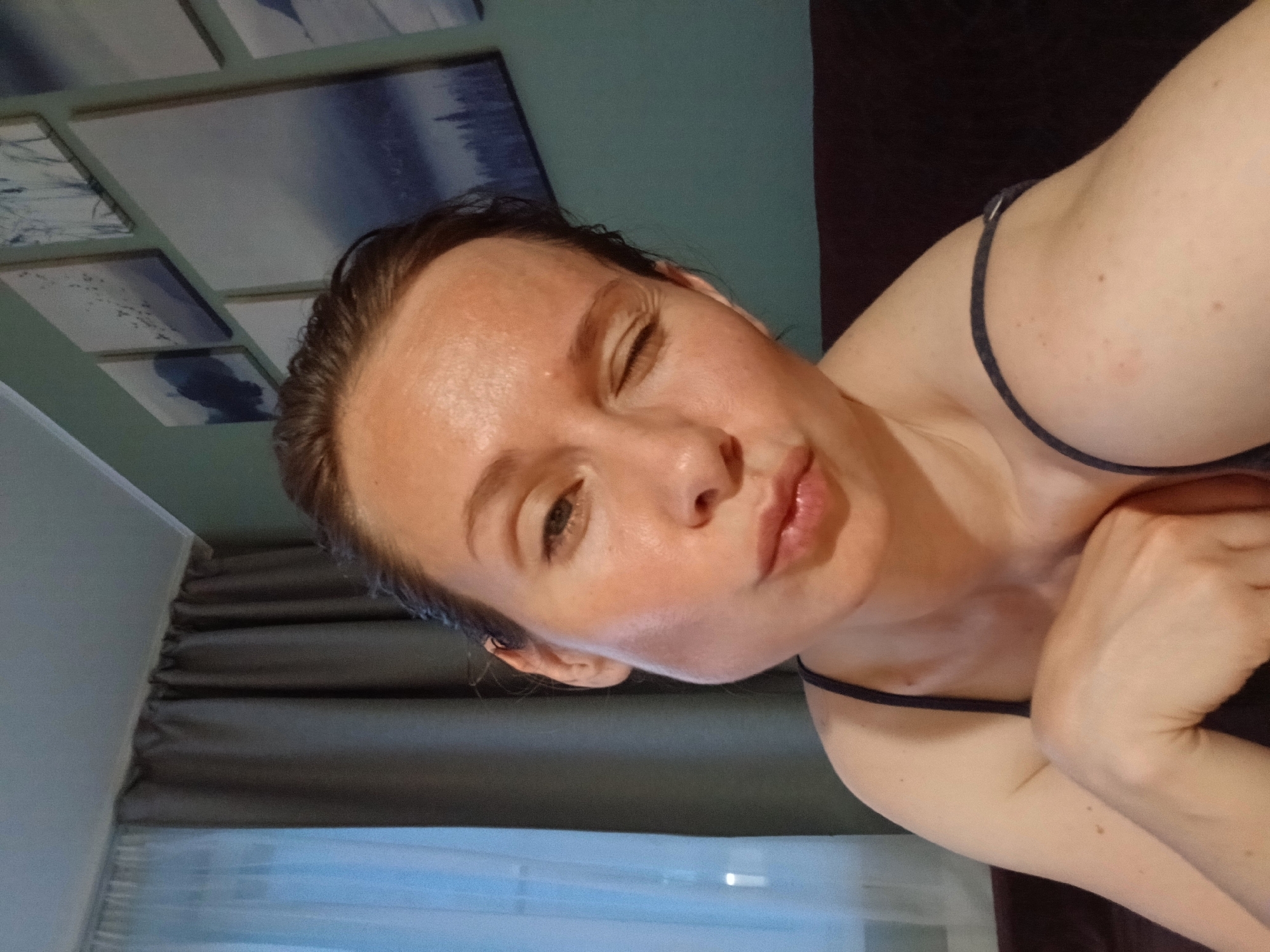 The image is a close-up selfie of a woman with her right eye closed in a wink and her lips puckered. She is wearing a dark camisole. The background shows a wall with several framed abstract art pieces depicting landscapes. To the left, there are curtains, one sheer and light blue, the other opaque and dark grey. The lighting suggests an indoor setting, possibly a home. There is no visible indication of the city of Yekaterinburg, Russia, in the image.
