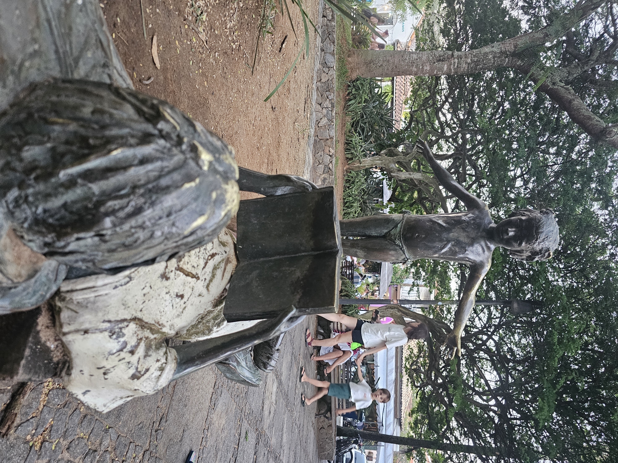 The image captures a scene in Armação dos Búzios, Brazil, featuring two bronze statues. One statue depicts a seated individual reading a book, positioned prominently in the foreground. The second statue, a standing figure with outstretched arms, is visible in the background. Between the statues, three children are interacting in the scene. The setting appears to be a public space with stone pavement and trees providing shade. The background includes glimpses of nearby buildings and structures, indicating an urban environment.