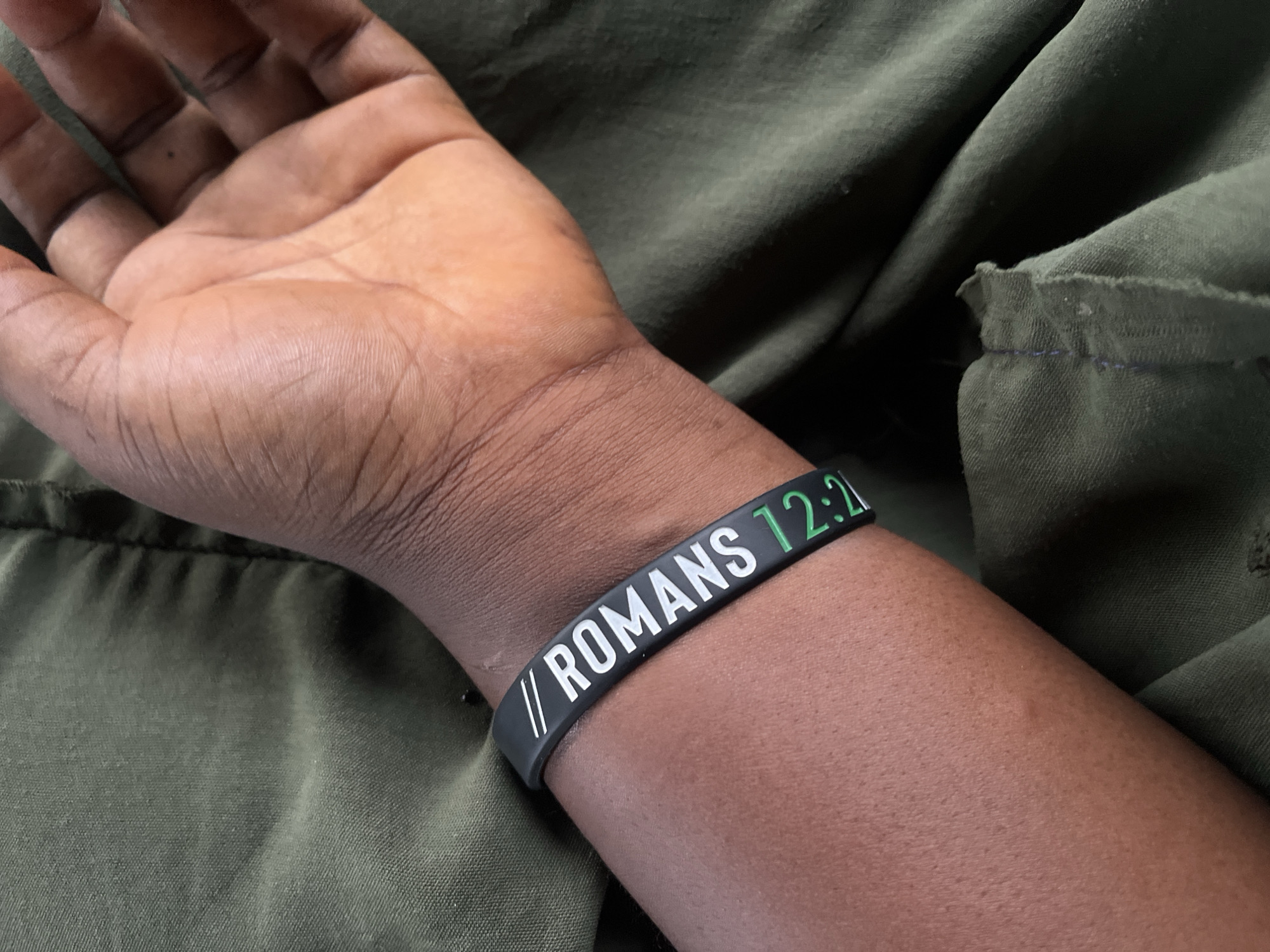 The image shows a close-up of a person's arm wearing a black silicone wristband with white text that reads "// ROMANS 12:2". The background is out of focus, showing dark green fabric. The lighting suggests an outdoor setting. There is no information in the image that would indicate its location in Jalingo, Nigeria.