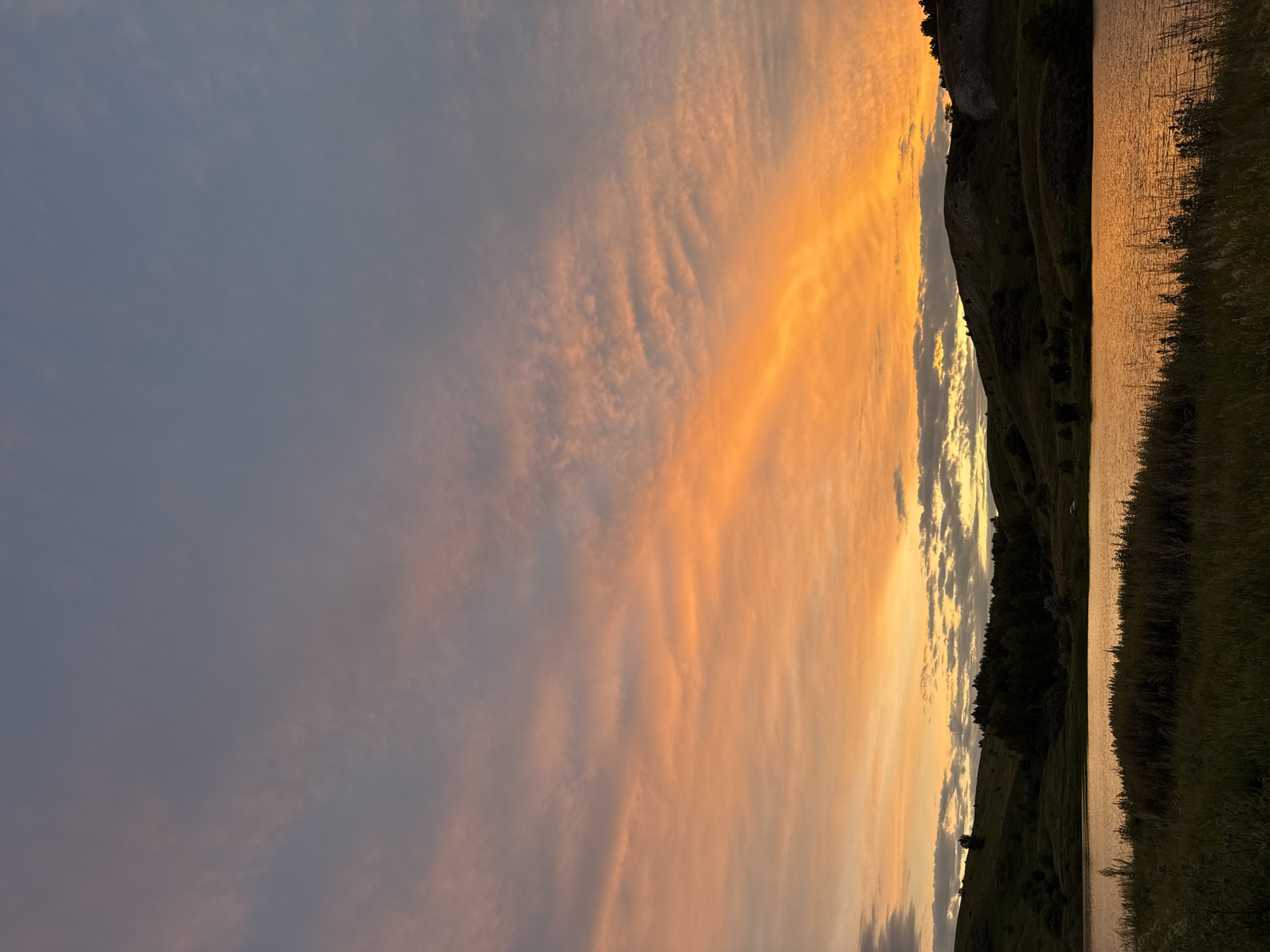 A scenic landscape captures a tranquil lake reflecting the warm hues of a sunset. Rolling hills dotted with trees form the backdrop, their dark silhouettes contrasting with the vibrant sky. The foreground features tall grasses and vegetation, adding texture to the scene. The sky is a canvas of soft blues and oranges, with wispy clouds painted across it, suggesting a calm evening in a natural setting.