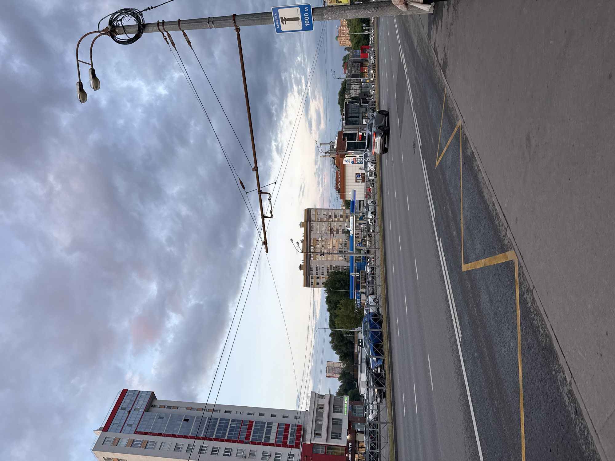 The image shows a street scene in Kazan, Russia, under a cloudy sky. In the foreground, a sidewalk with a yellow painted bus stop marking is visible. The street is a multi-lane road with white lane markings. On the left side of the street, a tall, modern building with red and white accents and many windows is prominent. Further down the street, there are other buildings, including a large apartment complex and a gas station with a blue awning. Several cars are parked on the side of the road and driving on the street. A lamppost with two lights and tangled wires stands on the right side of the image, displaying a blue sign with a white wrench symbol and "1000 M" text, indicating distance. The overall impression is an urban environment during daytime, possibly late afternoon or early evening given the soft lighting.