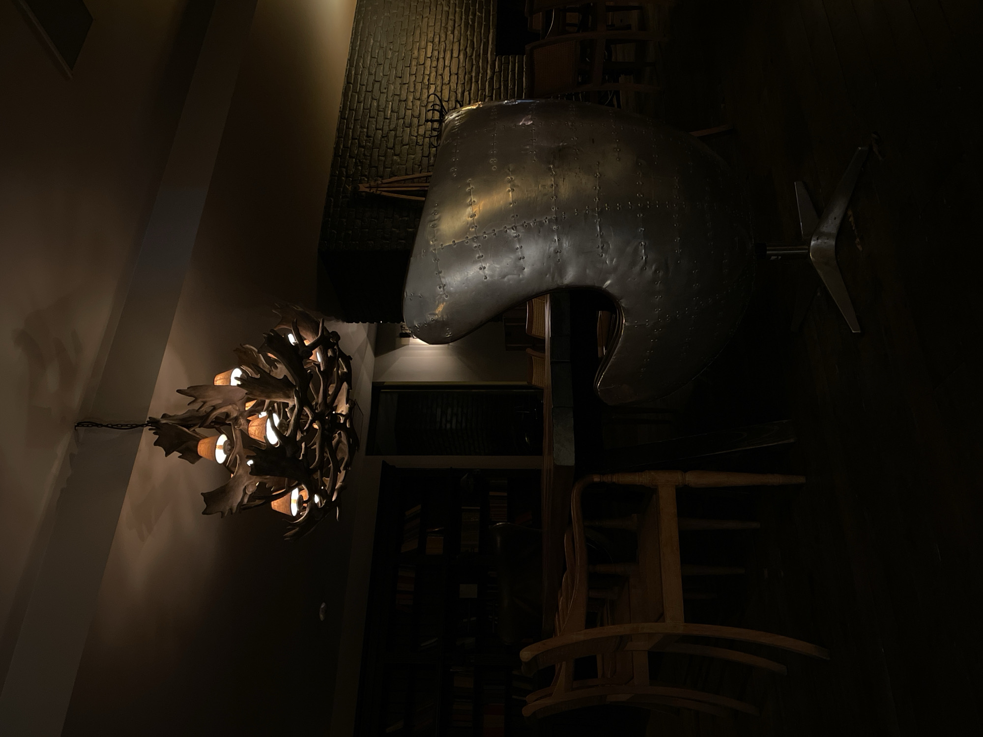 The image depicts an interior scene with a dimly lit ambiance, characteristic of a rustic or vintage establishment. Dominating the foreground is a large, metallic, egg-shaped chair with rivets, resting on a five-pointed metal base. Behind it, a long wooden table is surrounded by several wooden chairs with slatted backs. To the left, a tall bookshelf filled with books spans the wall. Above the table, a unique chandelier made of antlers and lit bulbs hangs from the ceiling. The background features a brick wall and, further back, other seating arrangements. The overall atmosphere is intimate and somewhat mysterious, enhanced by the low lighting and the combination of natural and industrial design elements. It's possible this scene is located in Stepantsminda, Georgia, a region known for its dramatic landscapes and opportunities for unique lodging.